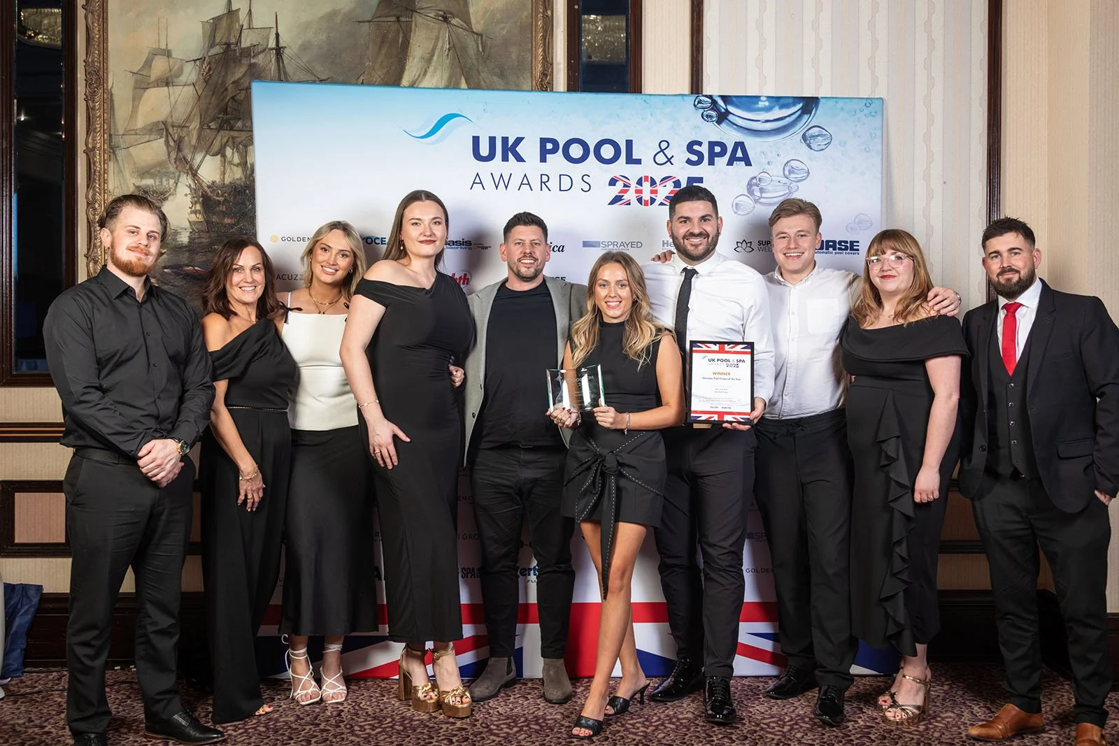 Group of eleven people dressed in formal attire, standing in front of a backdrop that reads 'UK Pool & Spa Awards 2022', celebrating an award win at a formal event.