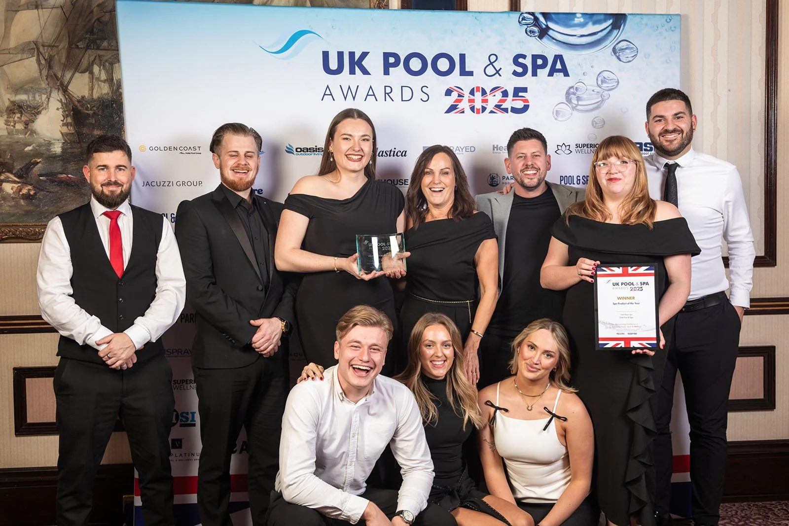 Group of eleven people, dressed formally, posing for a photo at the UK Pool & Spa Awards 2023 event. They are holding awards and certificates, standing in front of a backdrop with event branding and various sponsor logos.