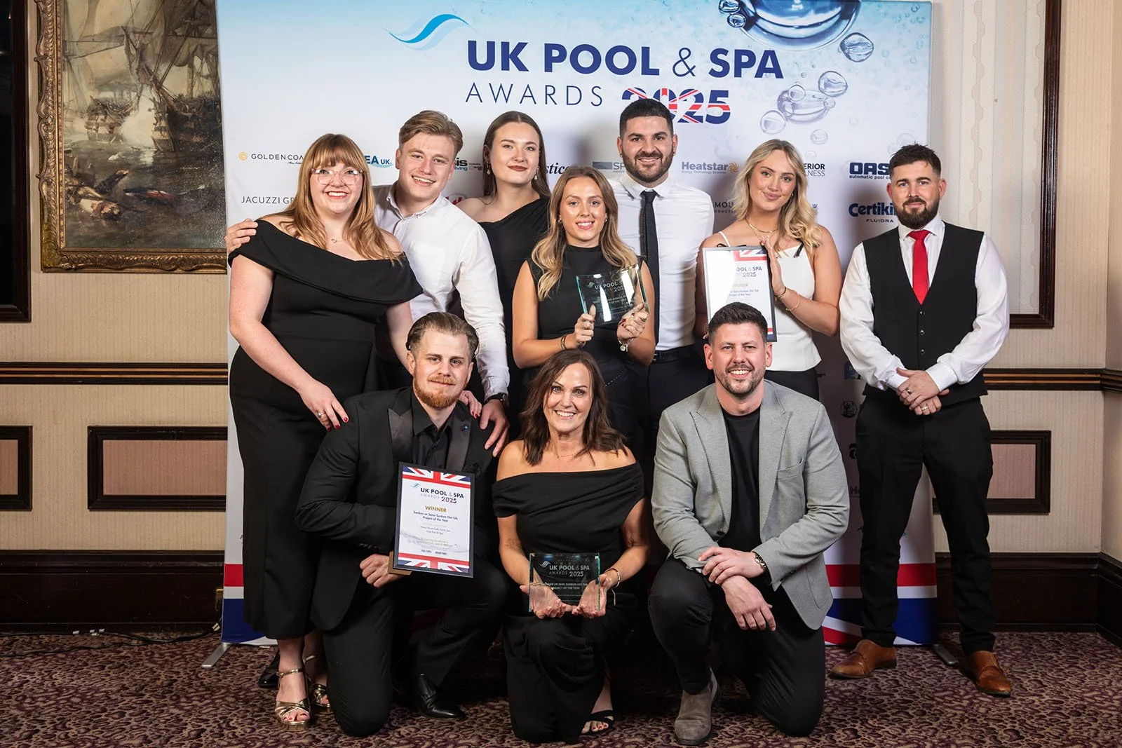 Group of people at UK Pool & Spa Awards 2023, some holding awards and certificates, posing in front of a banner with event details and sponsor logos, in a decorated indoor setting.