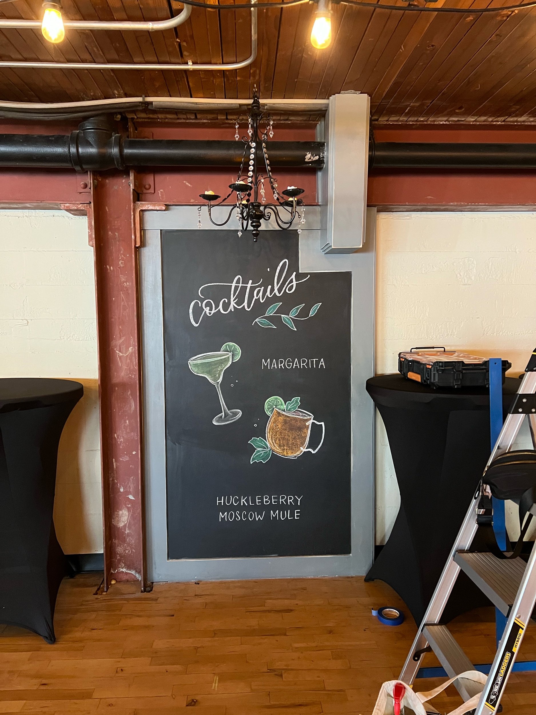 Chalkboard menu displaying "Cocktails" with illustrations of a margarita and a huckleberry Moscow mule, set in a rustic interior with a chandelier and step ladder nearby.