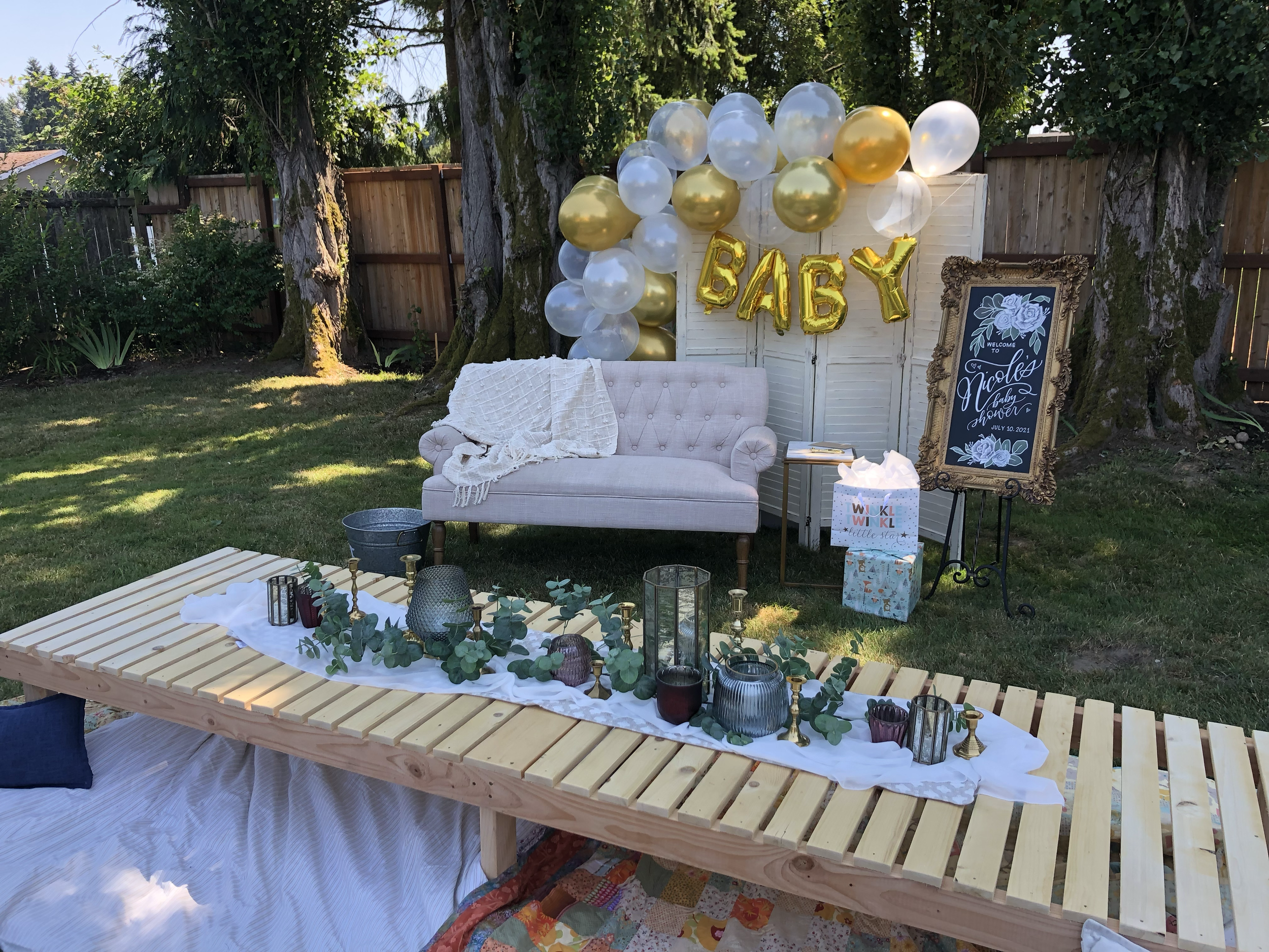 Outdoor baby shower setup with a white couch and gold and white balloons spelling 'BABY.' A table with candles and eucalyptus decor is in front, and a decorative sign stands to the side.