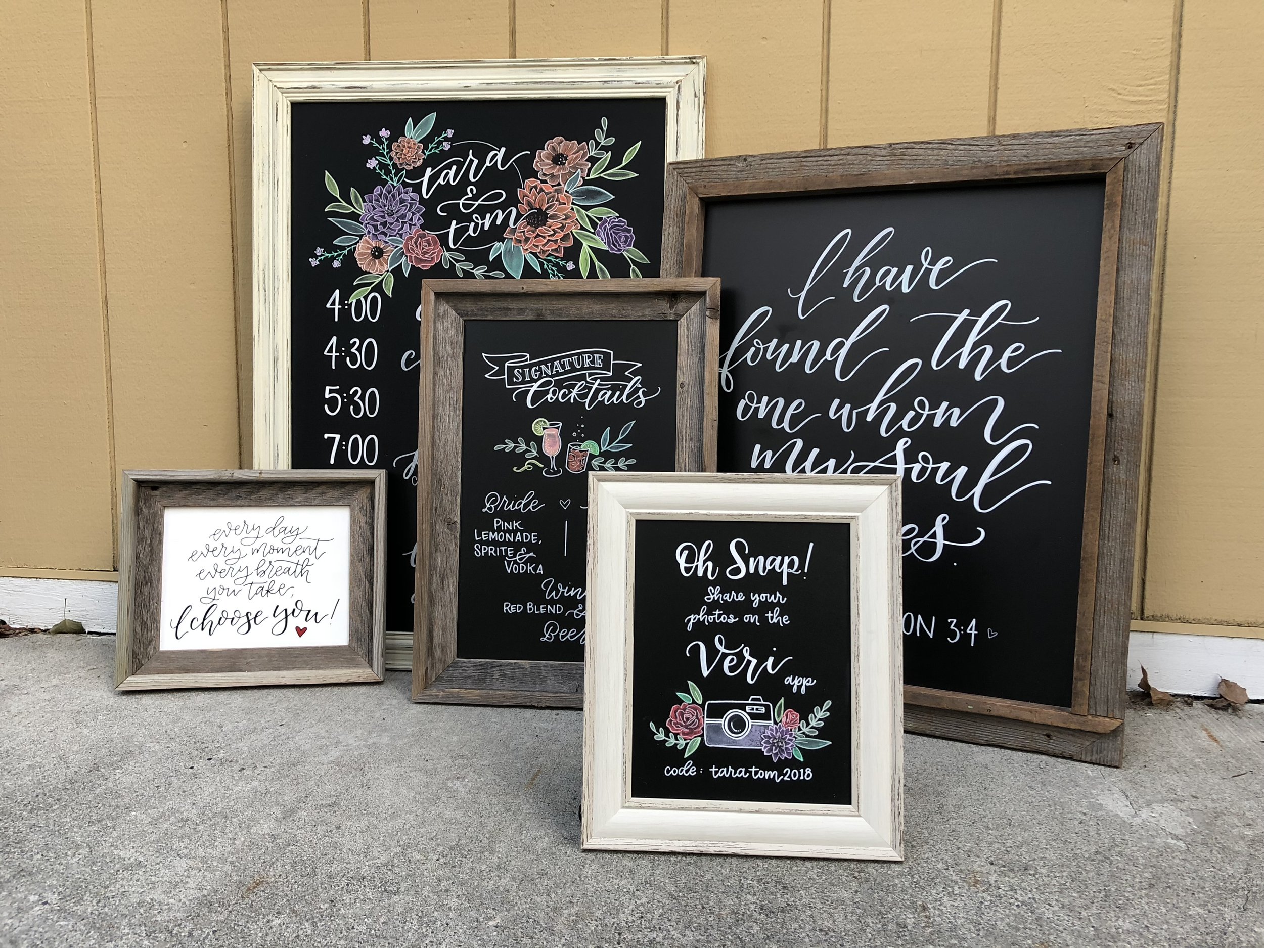 Collection of decorative framed wedding signs on the ground, featuring calligraphy and floral designs, including a schedule, cocktail menu, Bible verse, photo sharing instructions, and a romantic quote.