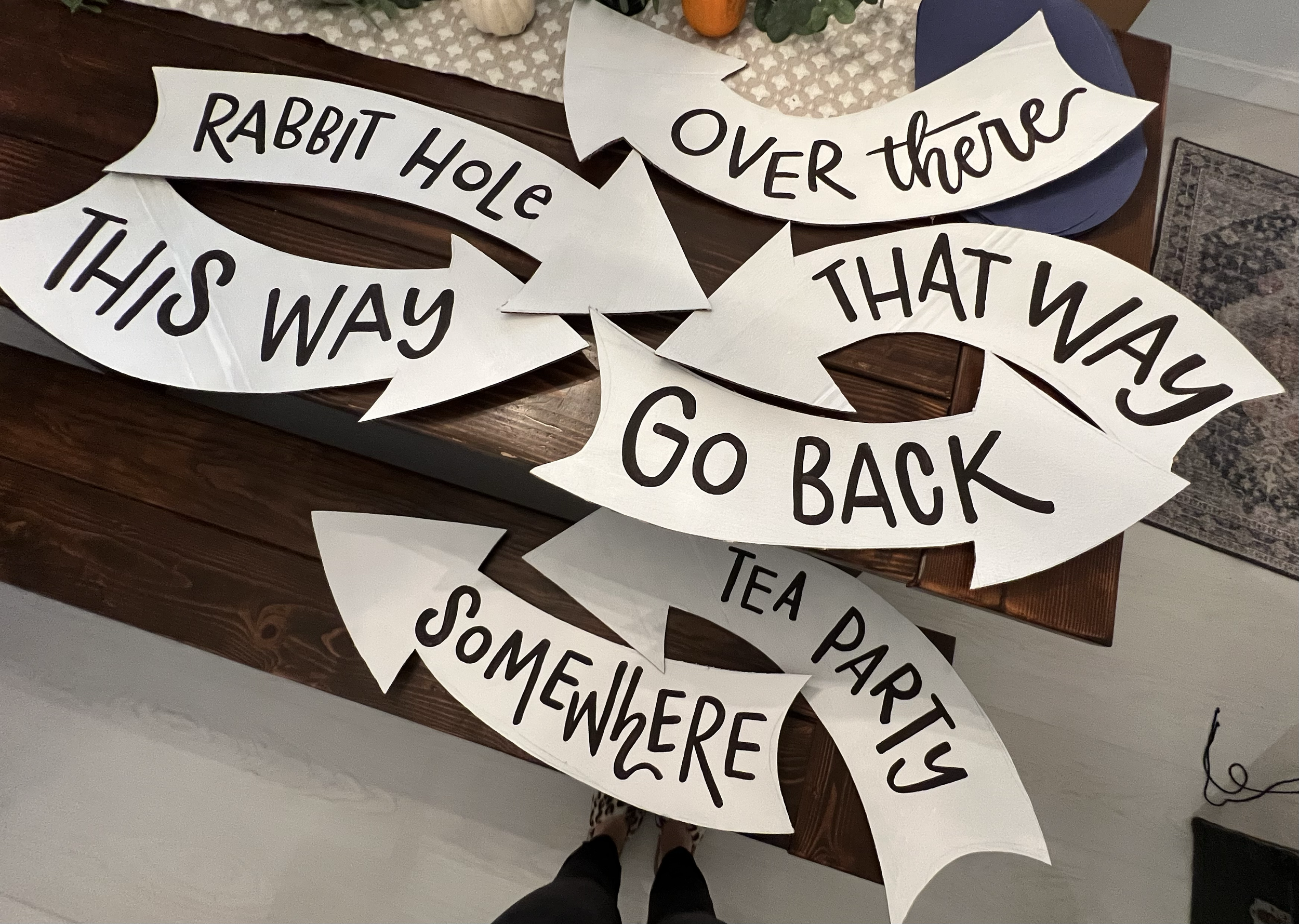Multiple white paper arrow signs with phrases, including "Rabbit Hole," "Over there," "This way," "That way," "Go back," "Somewhere," and "Tea Party," displayed on a wooden table.