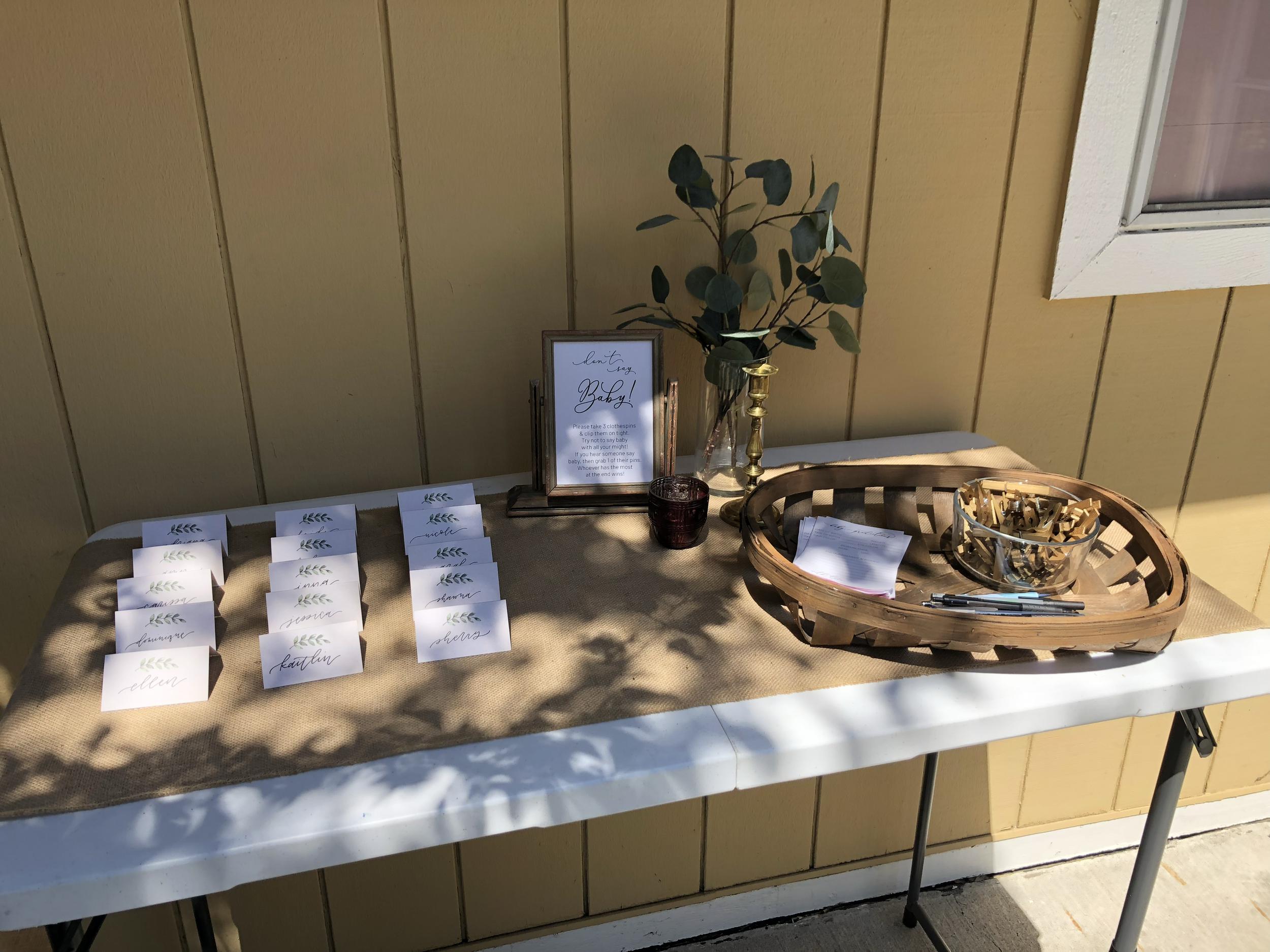 Table with name cards, a framed sign, greenery, a candle, and a basket with clothespins on a light tan tablecloth against a yellow wall.