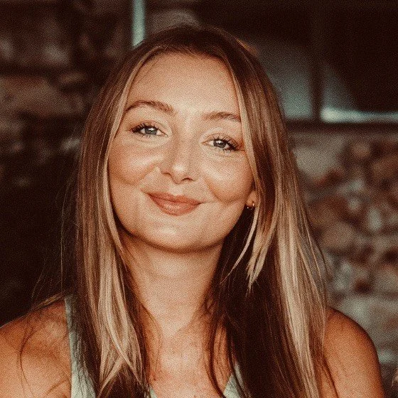 Close-up of a young woman with long light brown hair, smiling softly, wearing a sleeveless top, indoors with a blurred background.