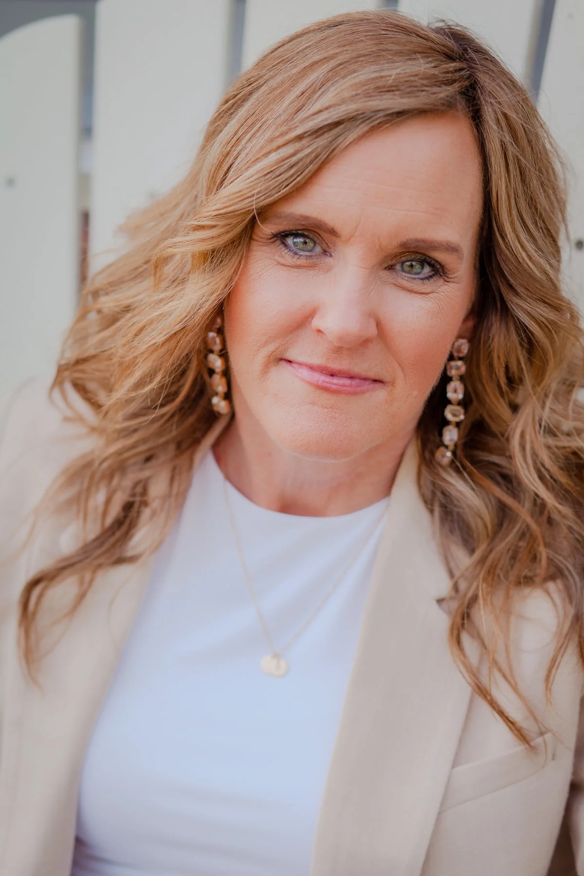 Close-up of a woman with wavy light brown hair, blue eyes, wearing a white top, beige blazer, gold necklace, and dangling earrings, outdoors near a white fence.