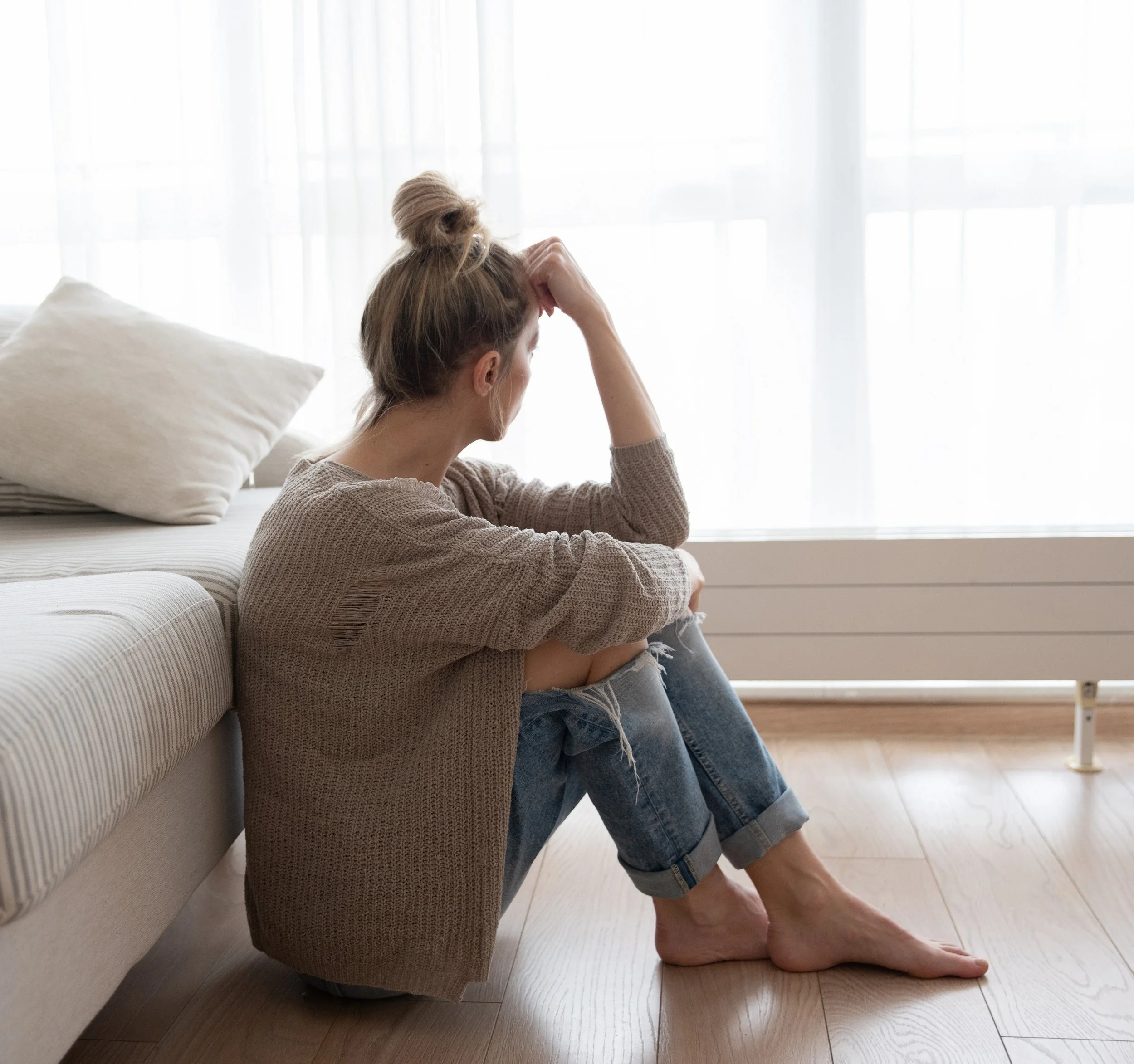 A woman sitting on the floor beside a couch, looking distressed, with her head resting in her hand, in a bright room with sheer curtains.