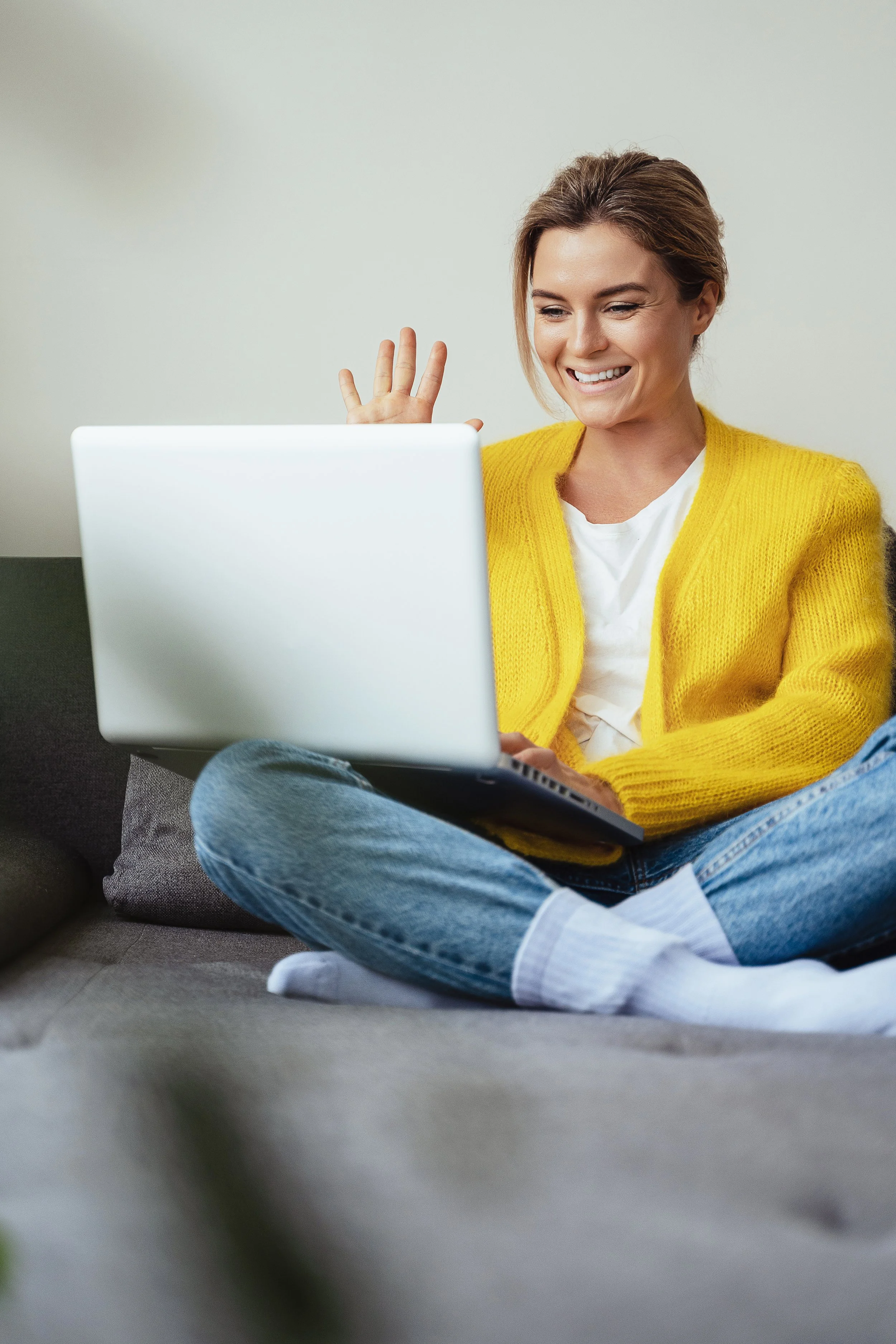 A woman sitting on a gray couch, smiling and waving at her laptop screen, wearing a yellow cardigan, white t-shirt, and blue jeans.
