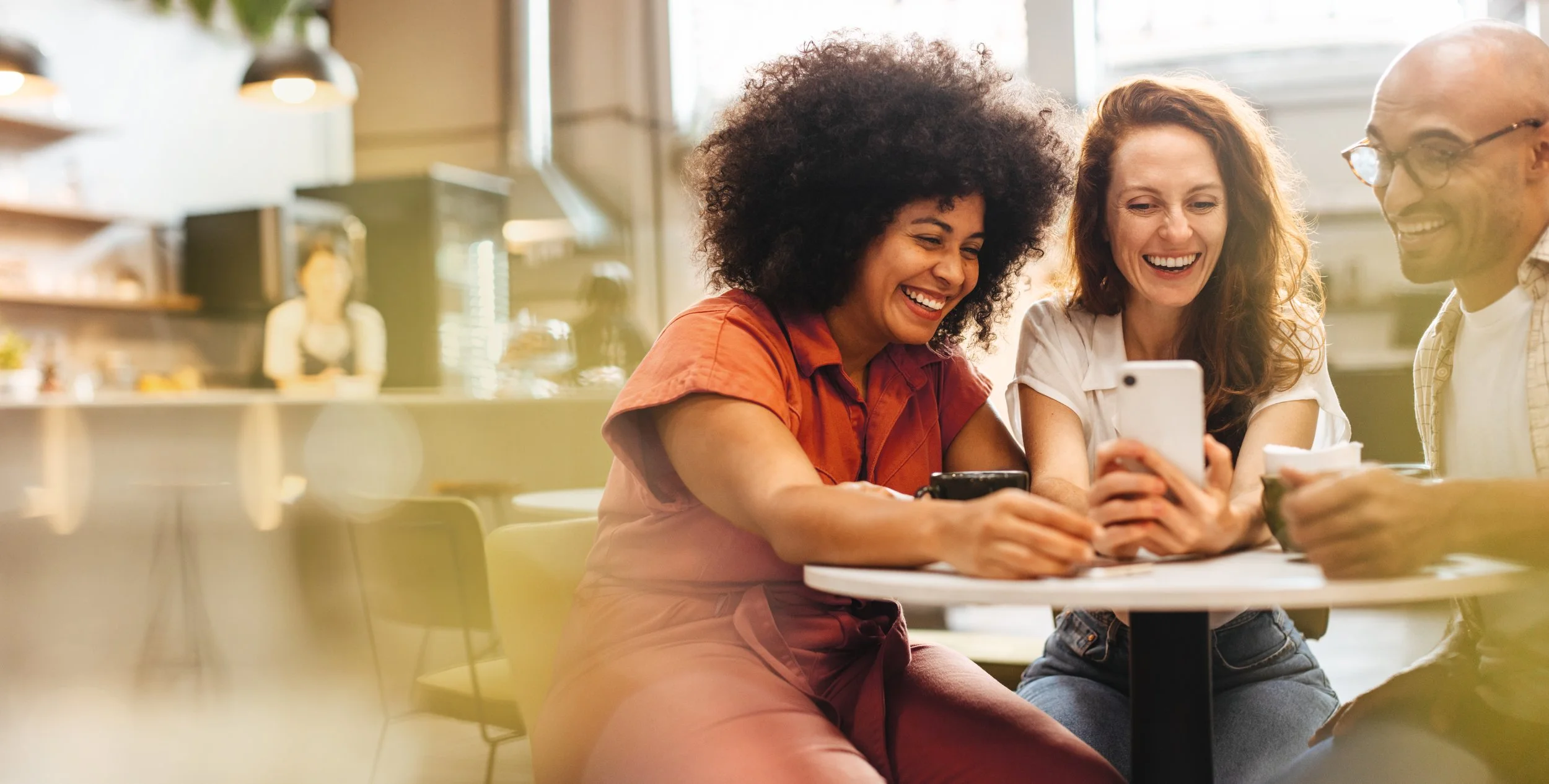 Three friends sitting at a table in a cafe, smiling and looking at a smartphone together.