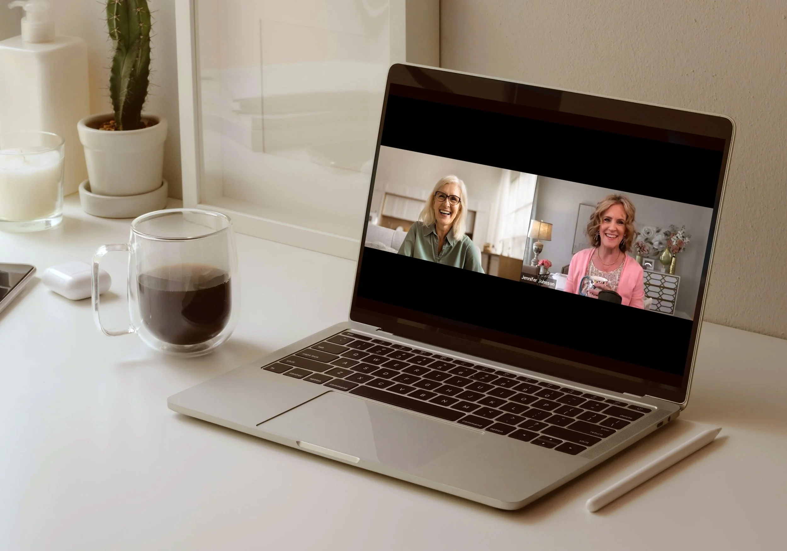 A laptop on a white desk shows two women in a video call, one with gray hair and glasses, and the other with curly blonde hair and a pink blazer.
