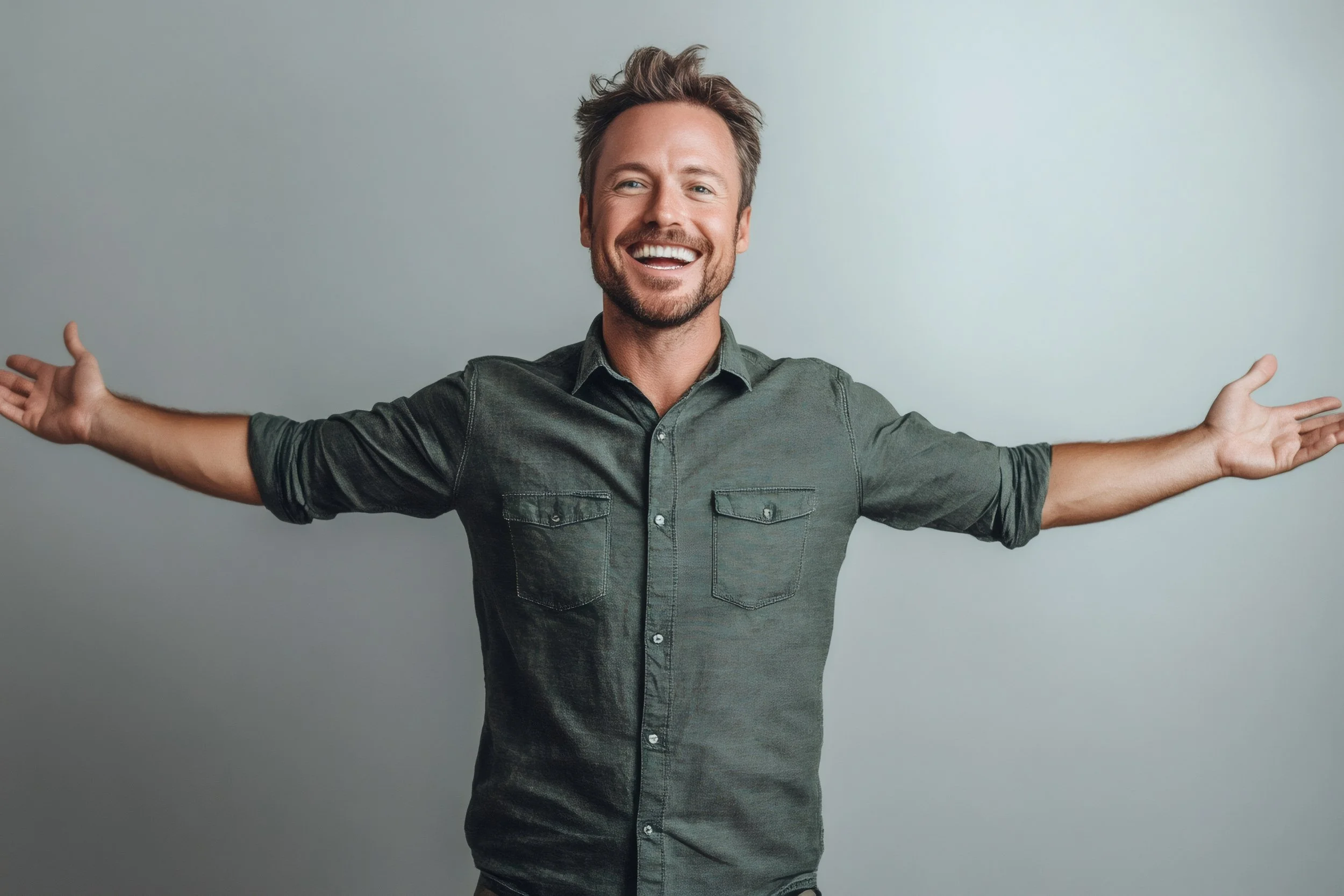 A smiling man with outstretched arms standing against a plain gray wall