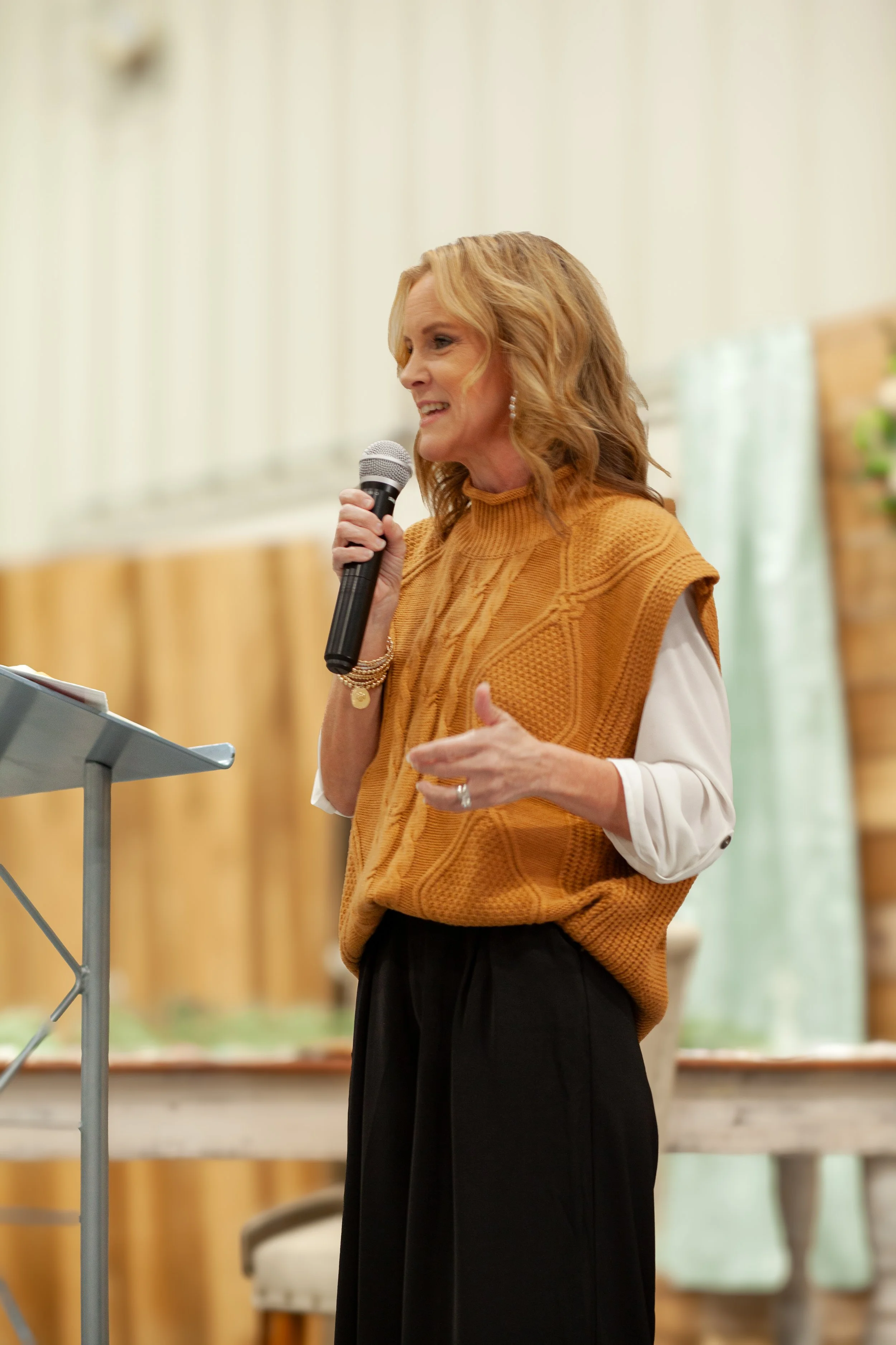A woman with shoulder-length red hair wearing a mustard yellow knit vest over a white blouse, holding a microphone, and speaking in a well-lit indoor setting.