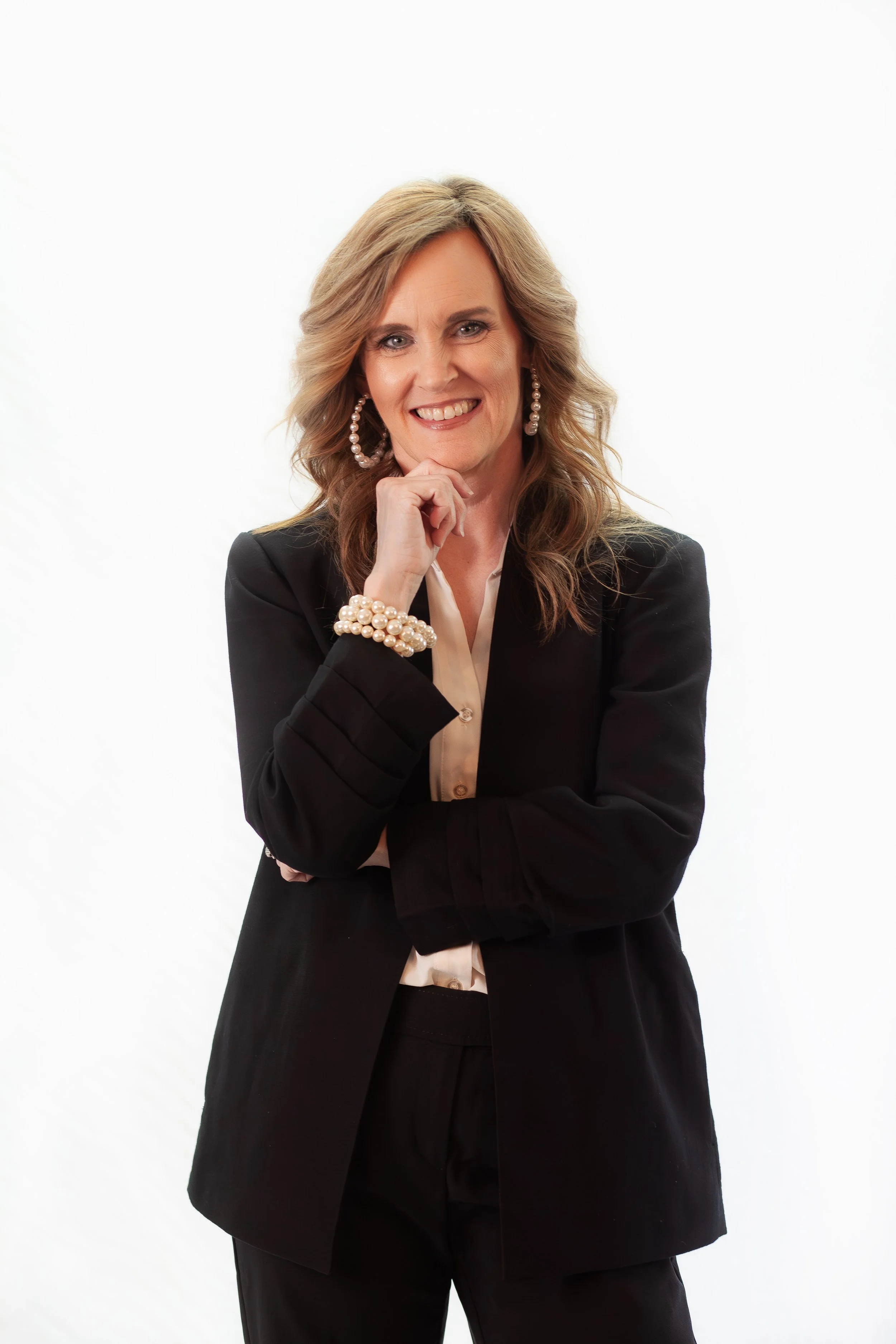 Smiling woman in black suit, pearl earrings, bracelet, and blouse, posing with hand under chin against a white background.