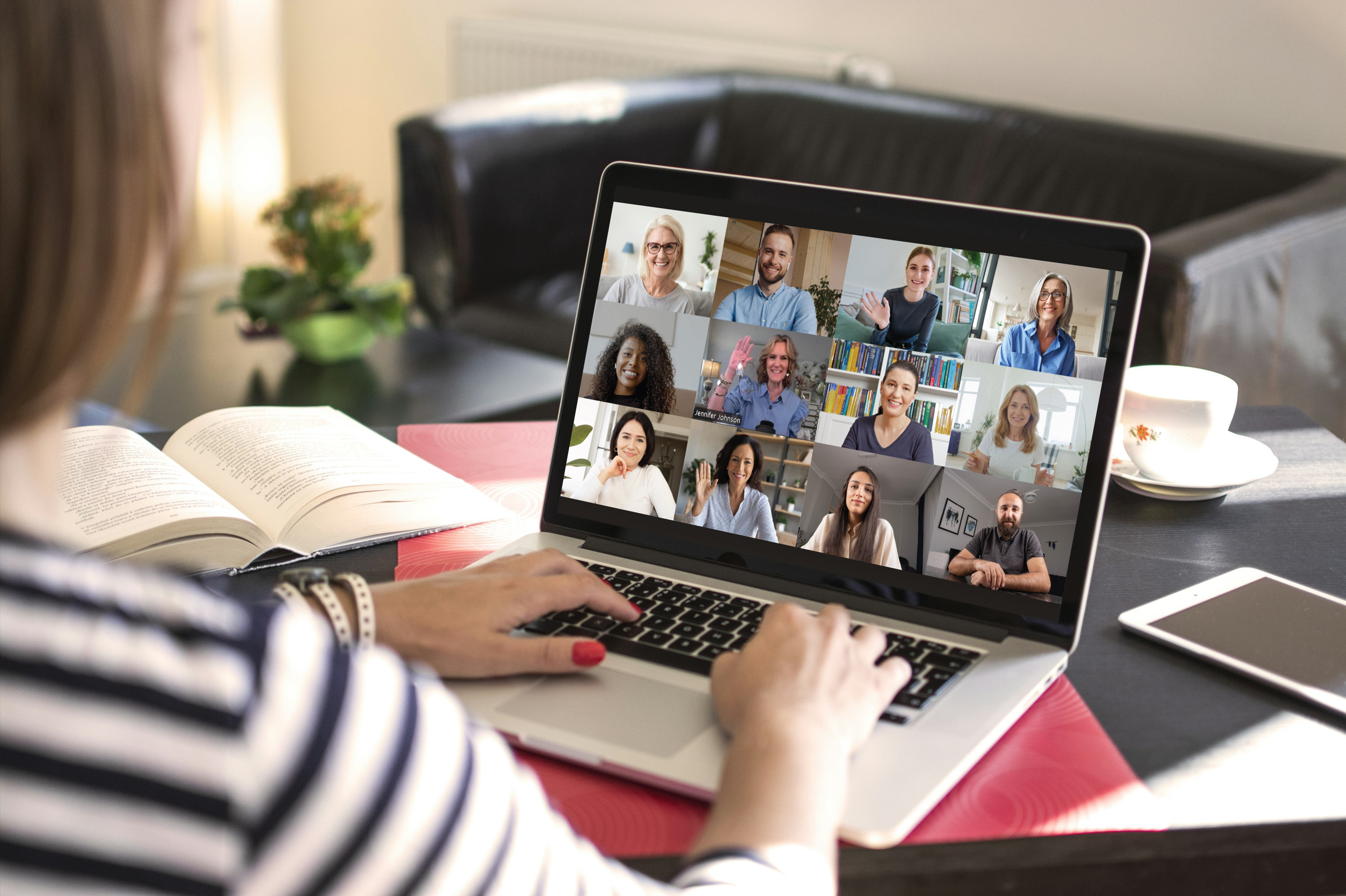 A woman with striped shirt practicing a virtual meeting on a laptop, with eleven smiling participants on the screen, in a cozy home setting.