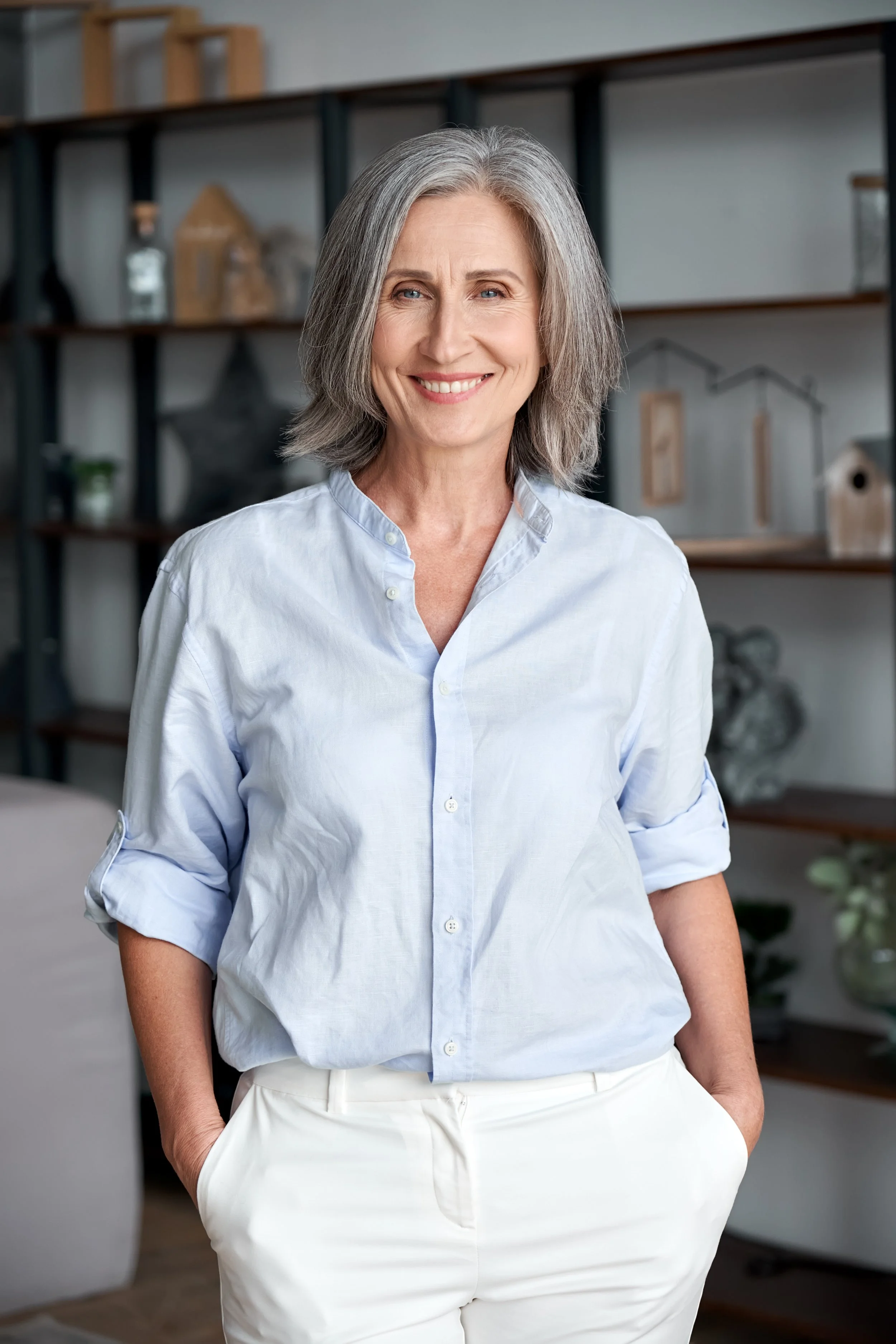 A smiling middle-aged woman with gray hair in a casual light blue button-down shirt and white pants, standing in a modern living room with shelves and decorative objects in the background.