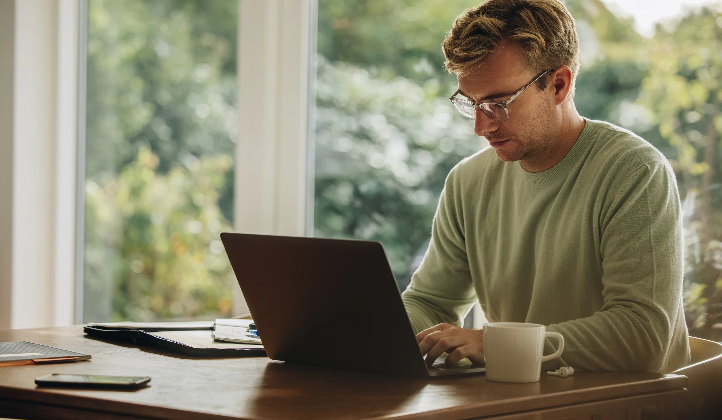 A young man with glasses, wearing a green long-sleeve shirt, working on a laptop at a wooden table inside near a window with a view of trees outside. There is a white mug, a notebook, a smartphone, and some papers on the table.