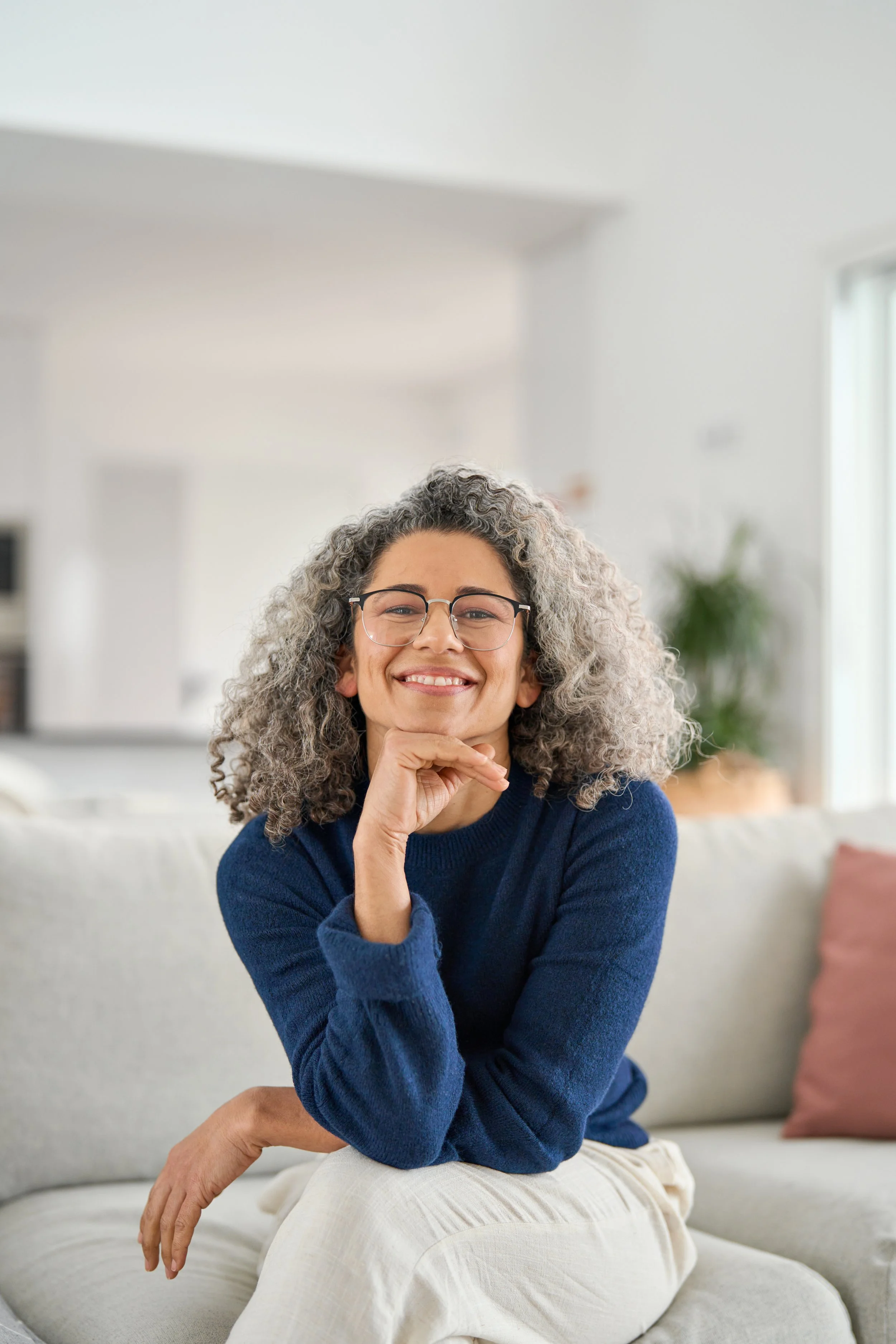 A smiling woman with curly gray hair and glasses, wearing a navy sweater, sitting on a light-colored sofa in a bright, modern living room.