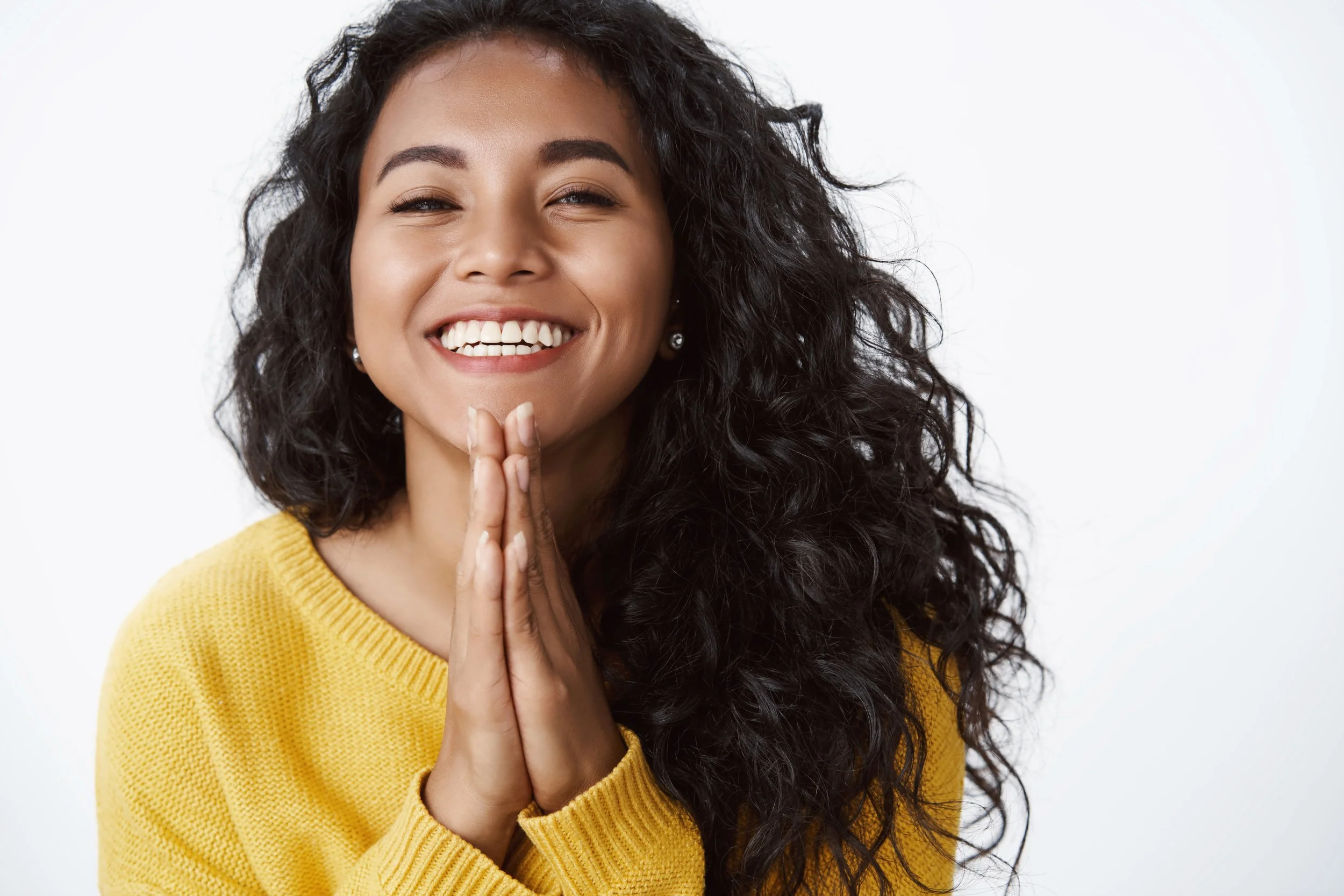 A woman smiling with hands pressed together in front of her face, wearing a yellow sweater and earrings, against a white background.