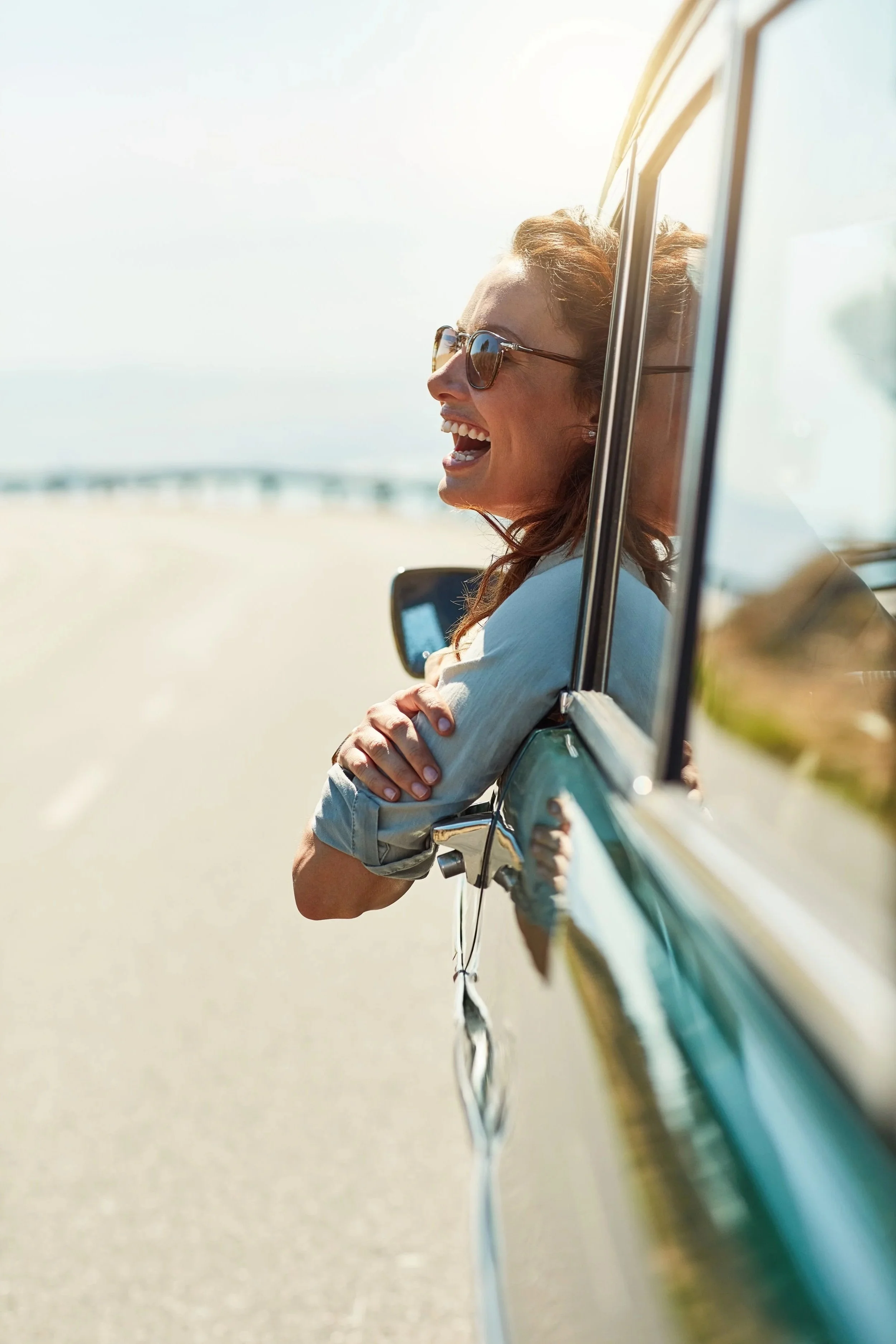 Woman wearing sunglasses smiling and laughing through open car window on a sunny day at the beach.
