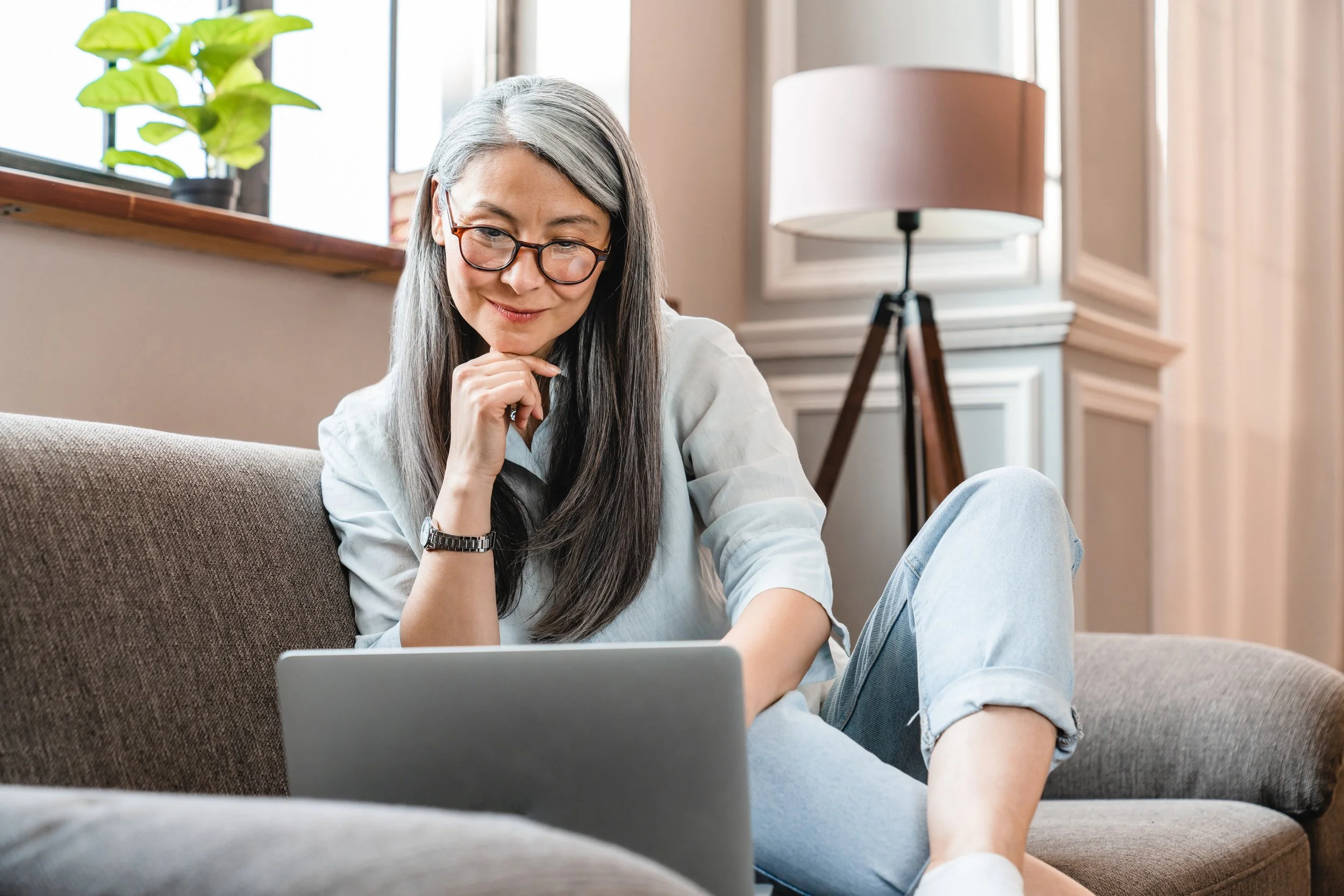 A woman with long gray hair and glasses sits on a beige sofa, looking at a laptop, in a cozy living room with a window, plant, and floor lamp.