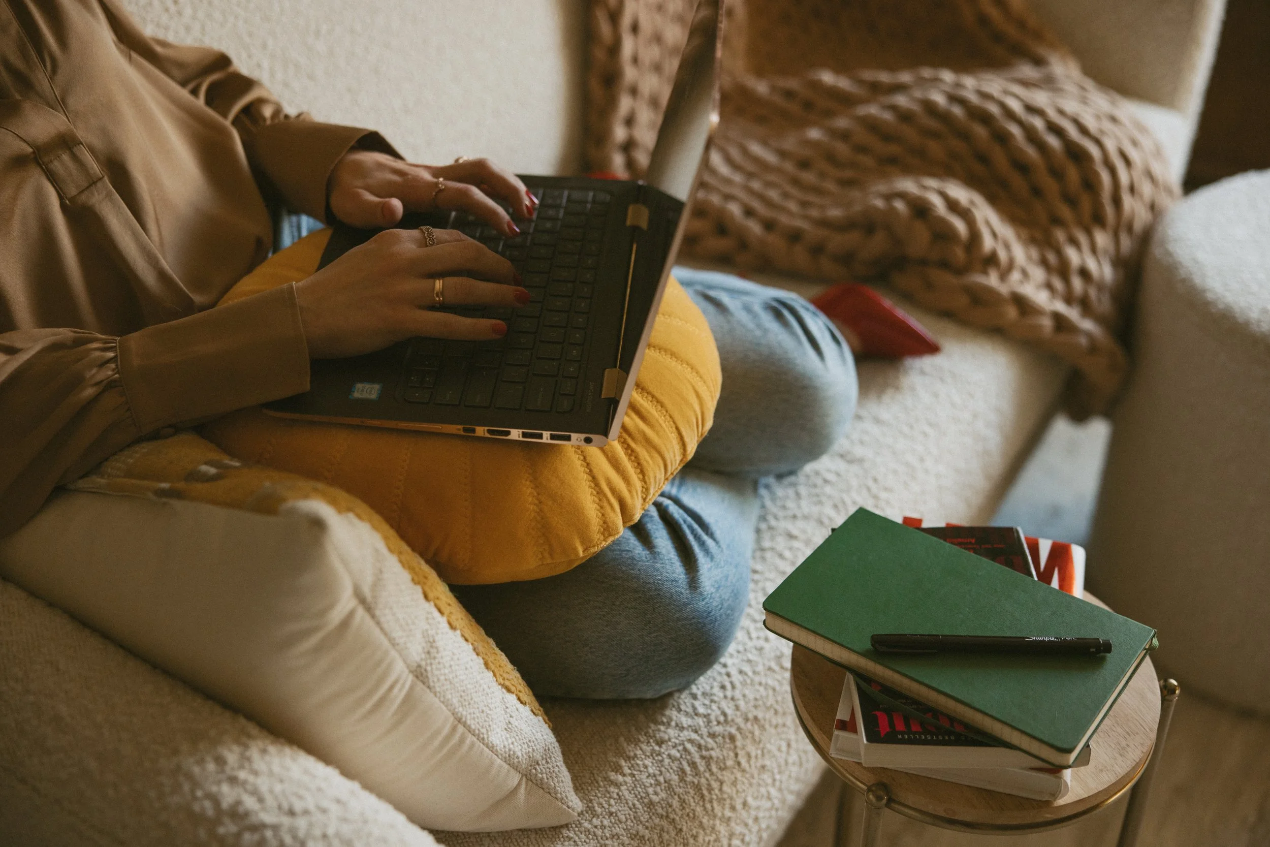 An anonymous woman sits with her feet curled up under her, typing on a laptop in her lap, surrounded by pillows, blanked and books.