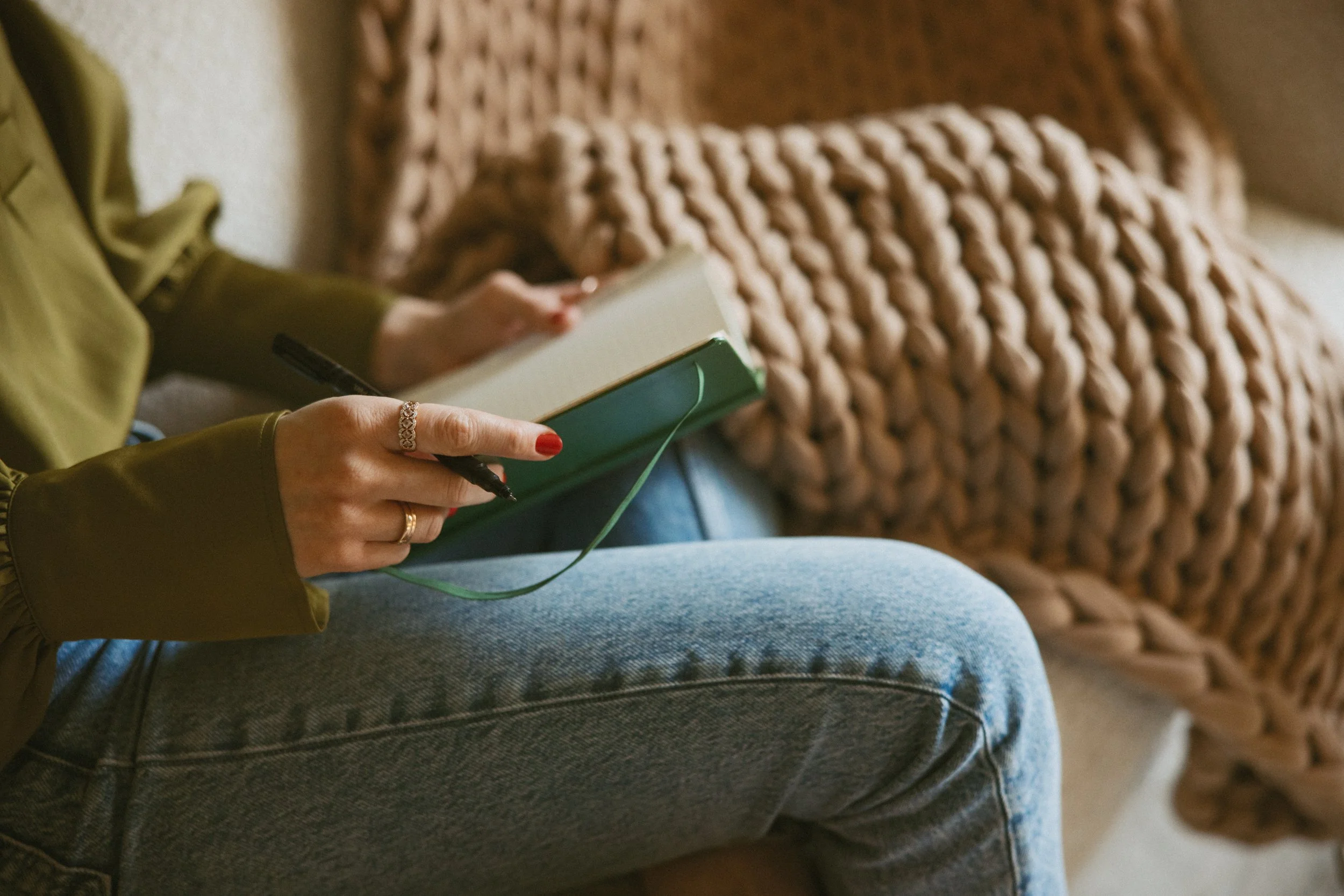Woman in green blouse and blue jeans holding a book next to a chunky tan blanket