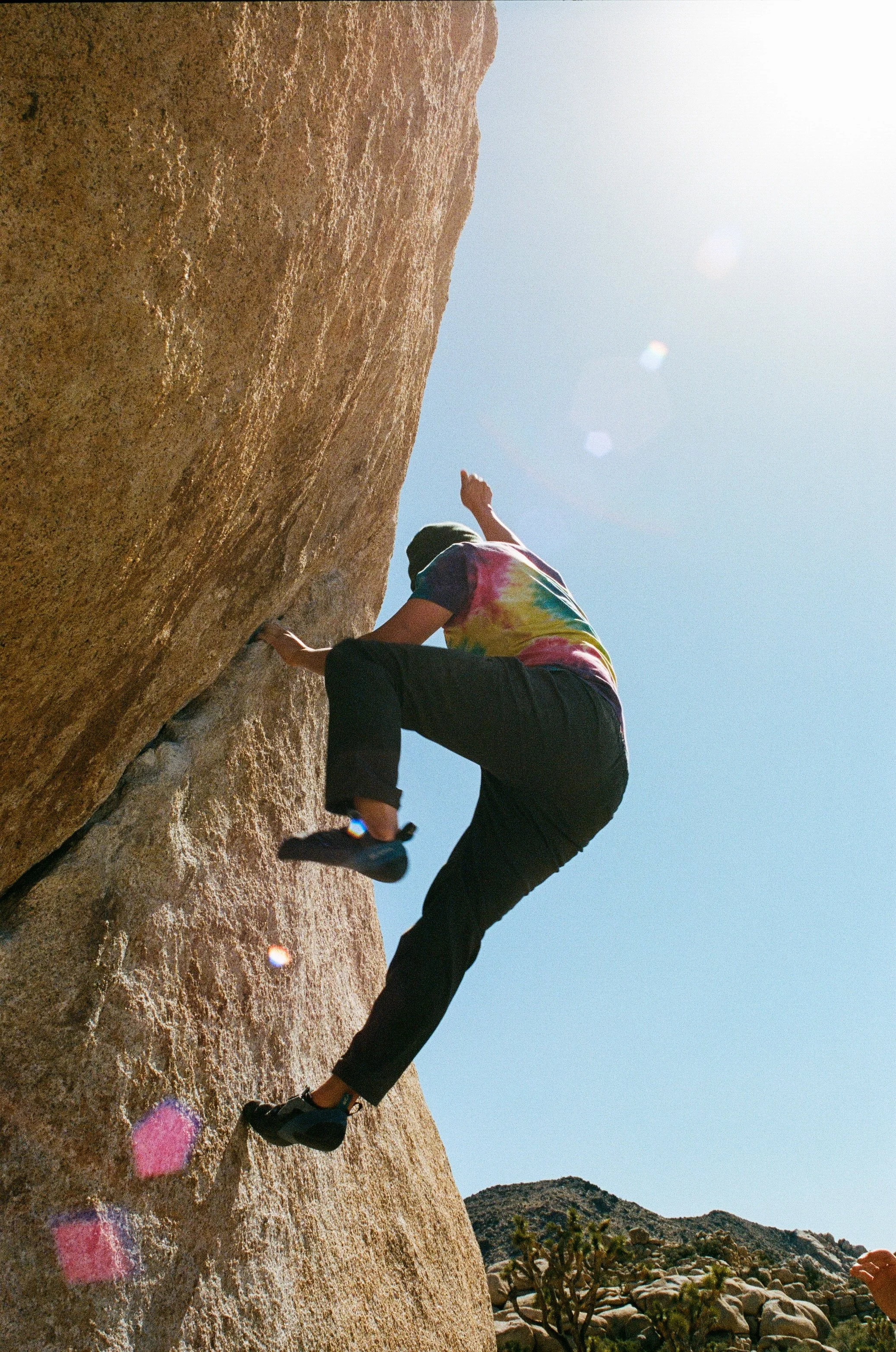 A person wearing a tie-dye t-shirt, black pants, and sneakers rock climbing on a large rock face outdoors under a bright sun.