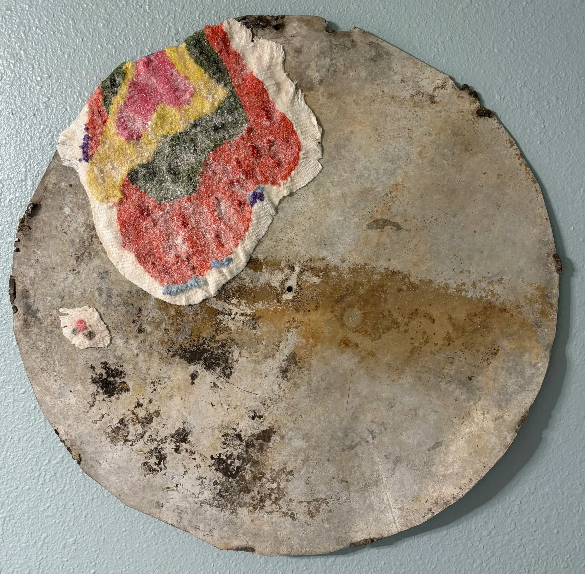 large round metal with a hand woven 'moon lichen' that is pink in the center, then green, then orange, then white, with some blue and purple for the large one sort of embracing the top left side of the metal with sculpted frayed edges + smaller 1