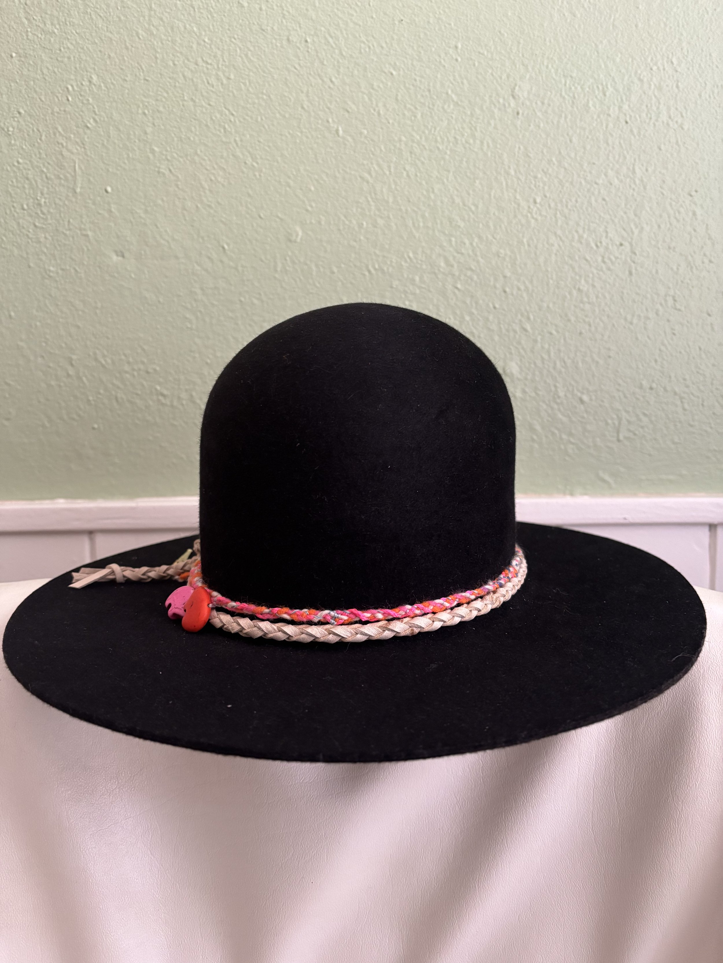 image of a black rounded hat with a light tan leather box-braided hat band layered with a multicolored rainbow and neon pink hat band with two zuni bear beads : a pink one and a red one.
