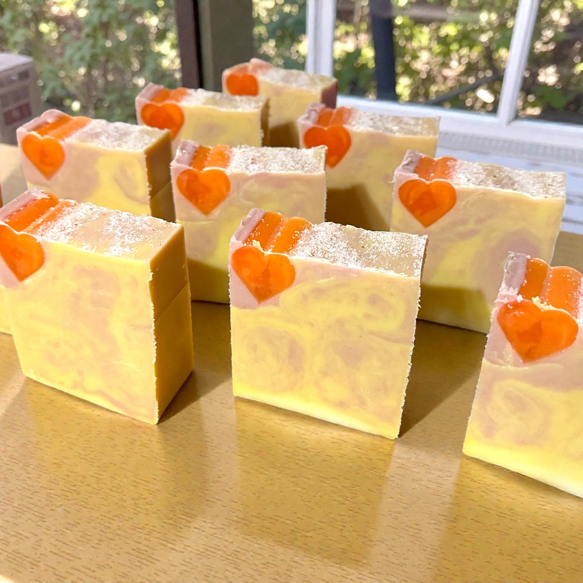 Multiple handmade soap bars with orange heart decorations on some top corners, arranged on a wooden surface, with a window and greenery in the background.