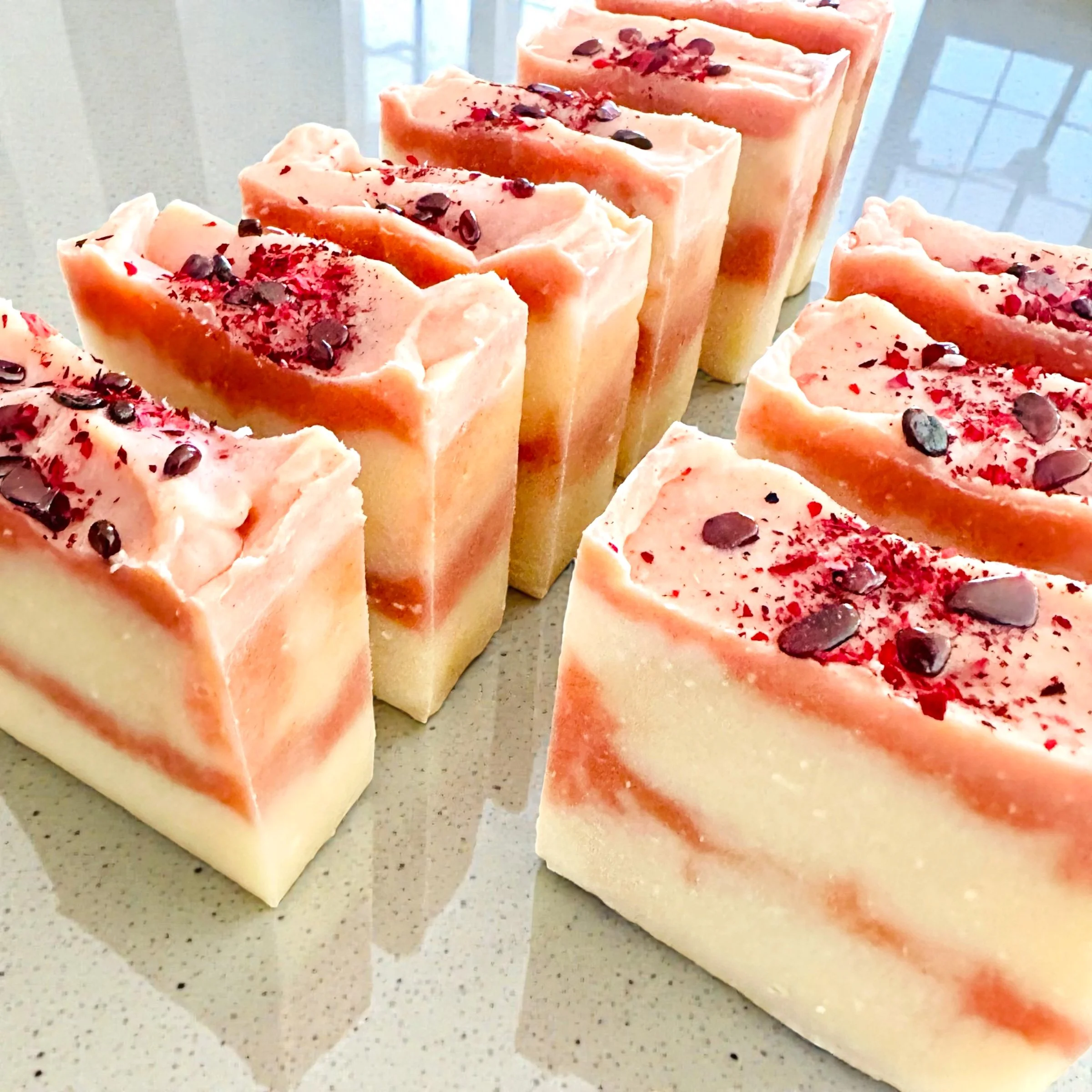 Slices of soap with layered pink and white colors, topped with red garnet chips and rose petals on a light-colored countertop.