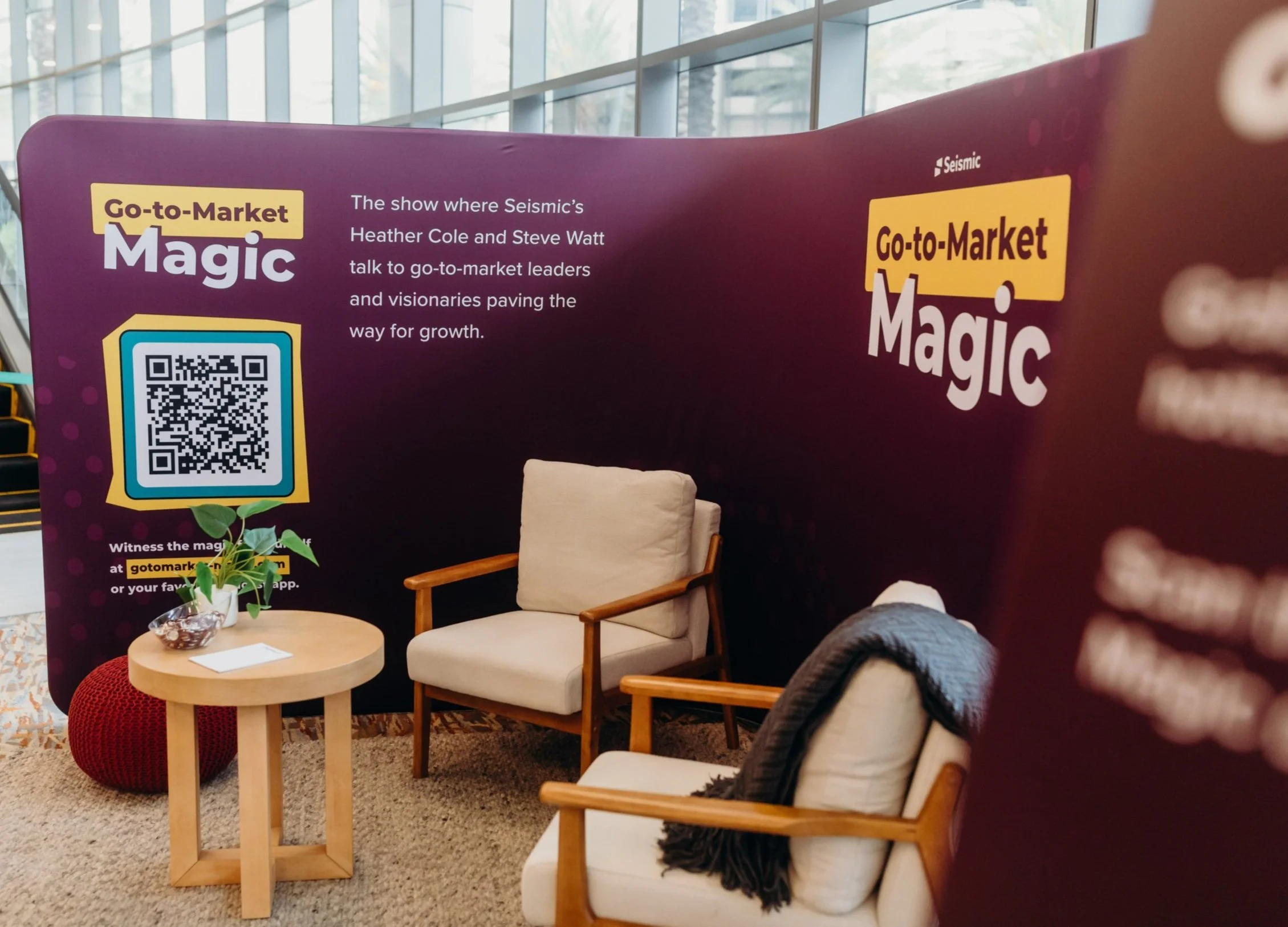 Event booth with purple backdrop displaying 'Go-to-Market Magic' and text about a podcast show by Seismic. Features chairs, a small table, and a QR code.