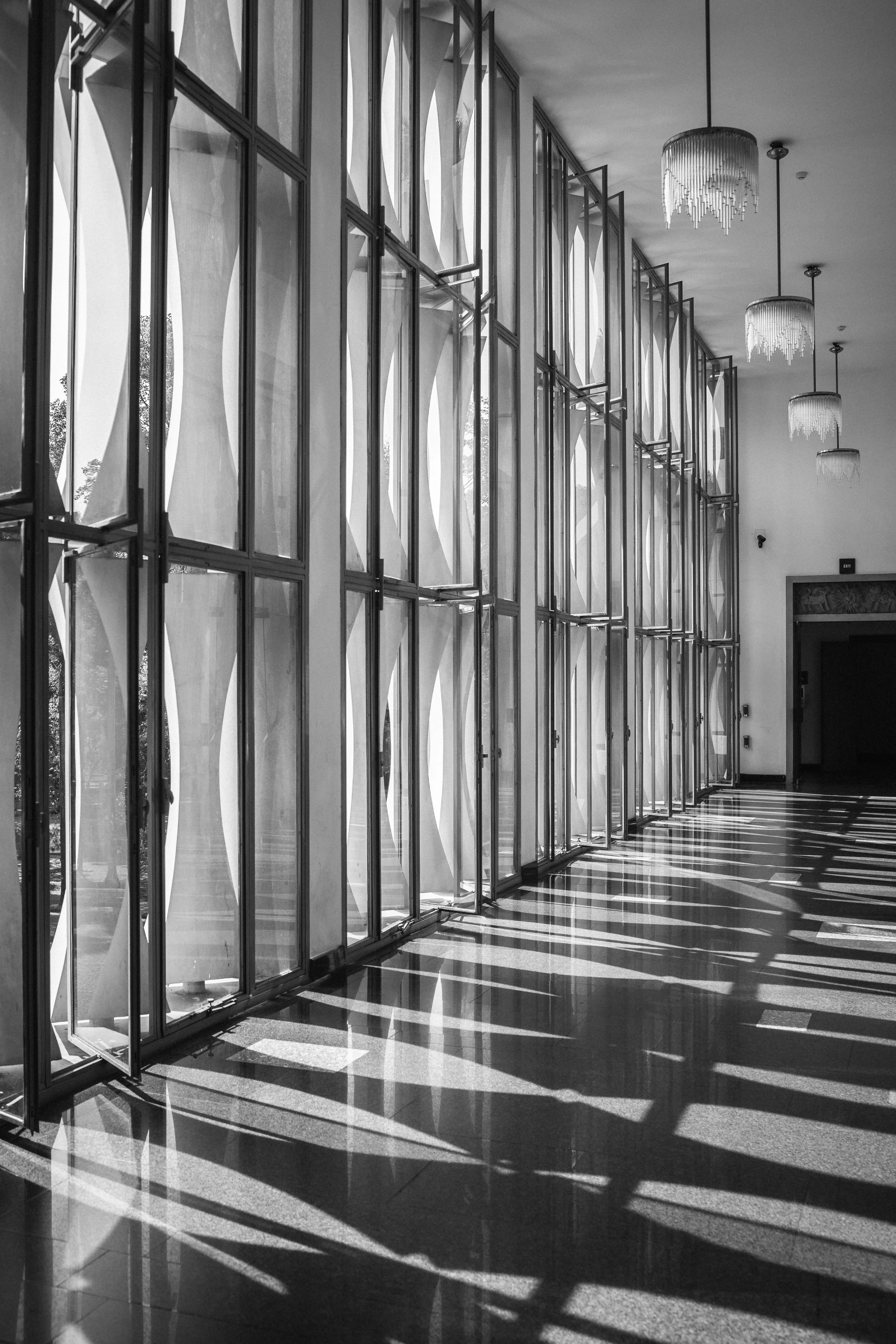 Sunlight casts shadows through a large window with multiple vertical panels in a modern interior space, with chandeliers hanging from the ceiling.