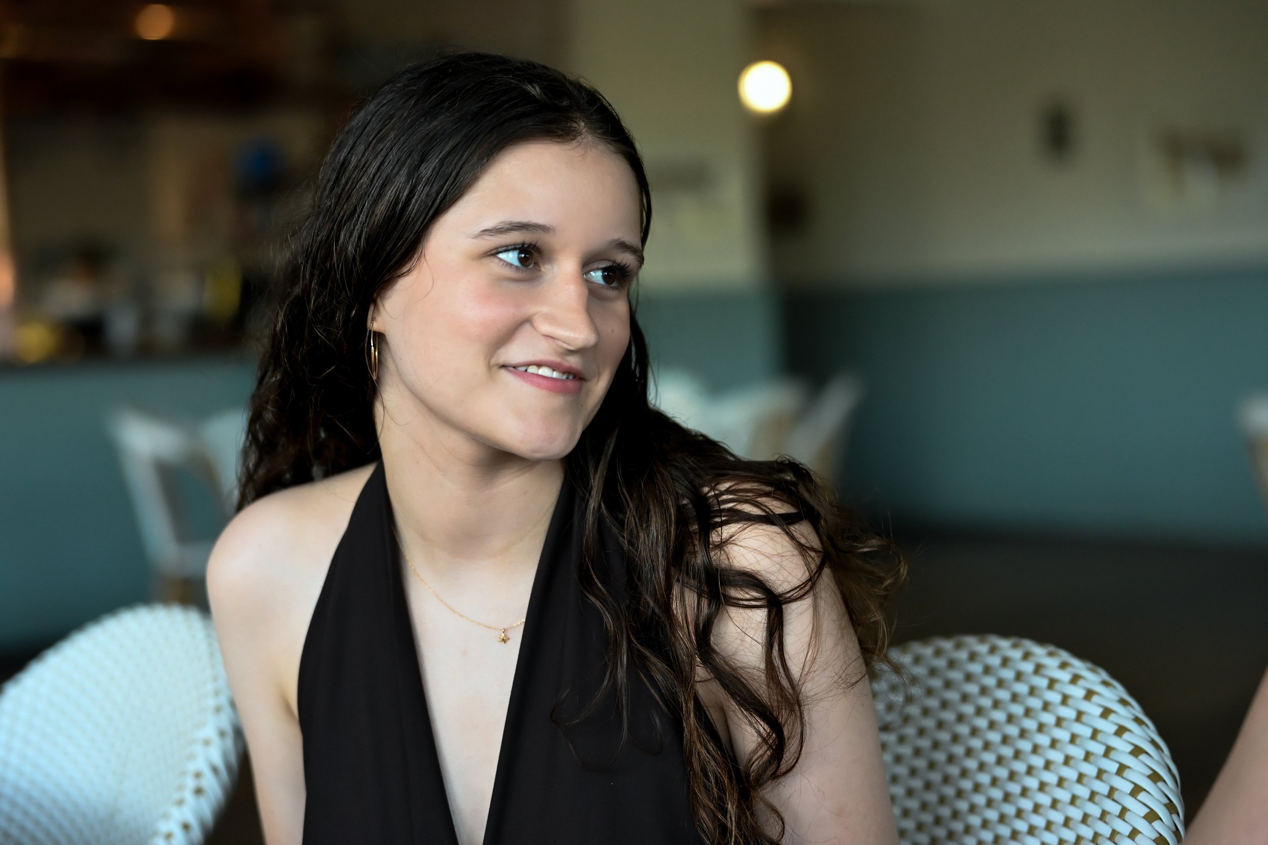 A young woman with long dark hair and brown eyes, wearing a black top, sitting in a cozy setting with modern decor, smiling slightly as she looks to her right.