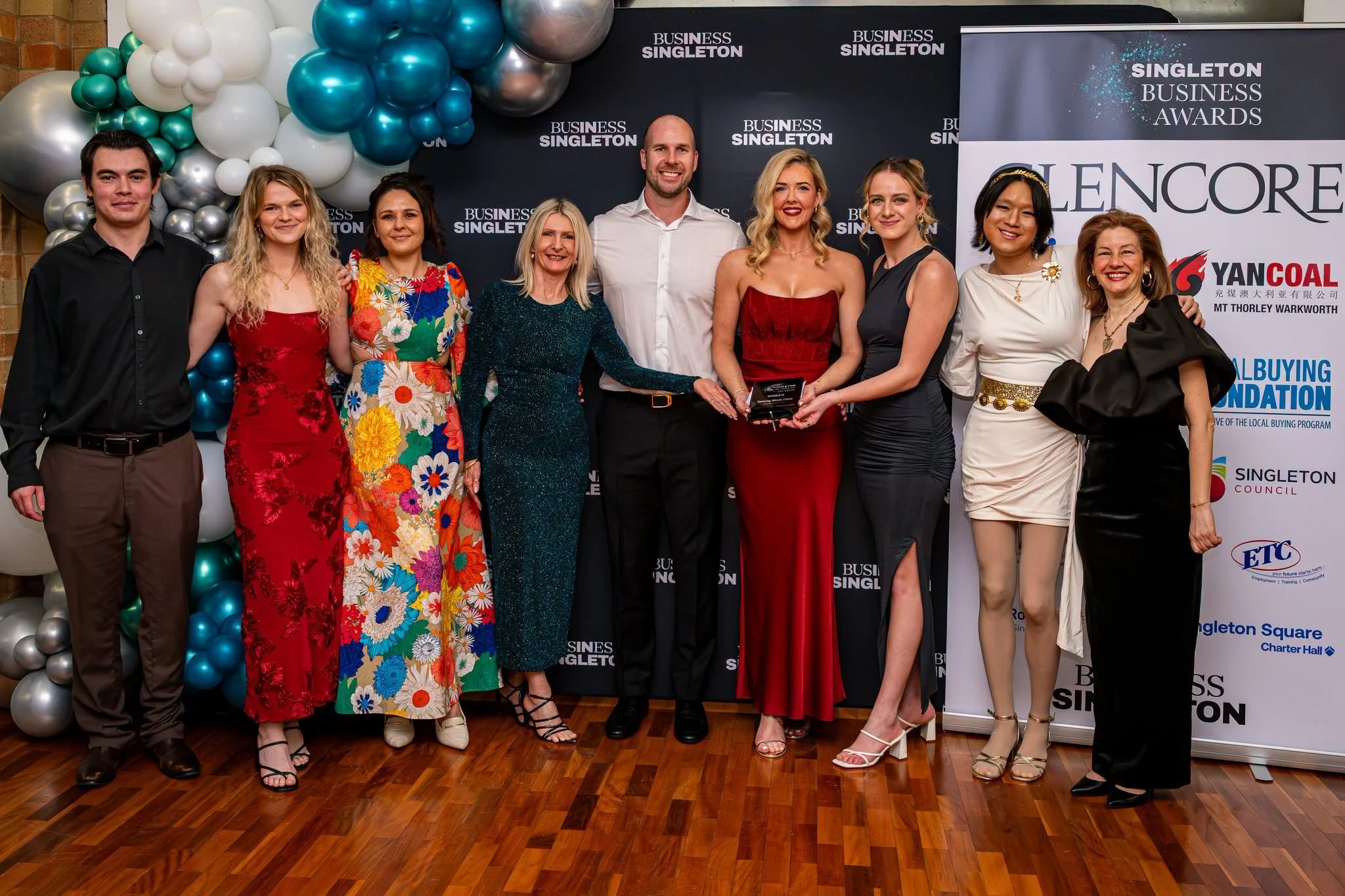 A group of ten diverse people standing together at a formal event, with some holding an award. Behind them are black banners with white text, a balloon arrangement of white, blue, and silver balloons, and some sponsor logos. The group is smiling and 