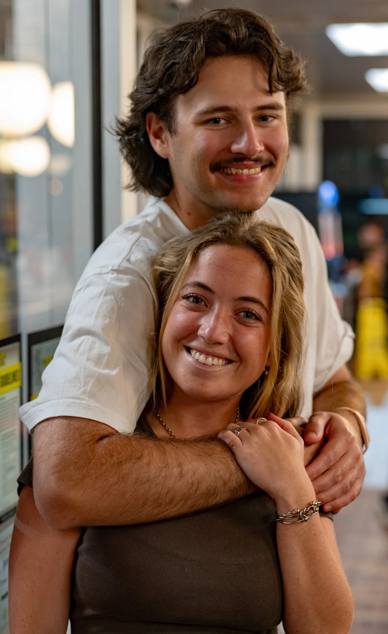 A young man and woman smiling in a friendly embrace at a restaurant or cafe with warm lighting.