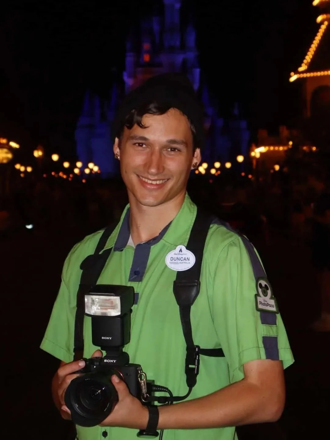 JOHN DUNCAN man in a green uniform holding a camera with a flash at a nighttime Disney park. Background has a lit castle and lights.