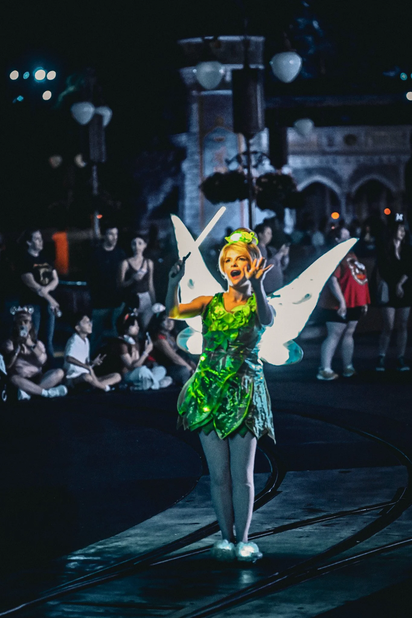 A performer in a green, butterfly-inspired costume on stage with an audience watching in the background.