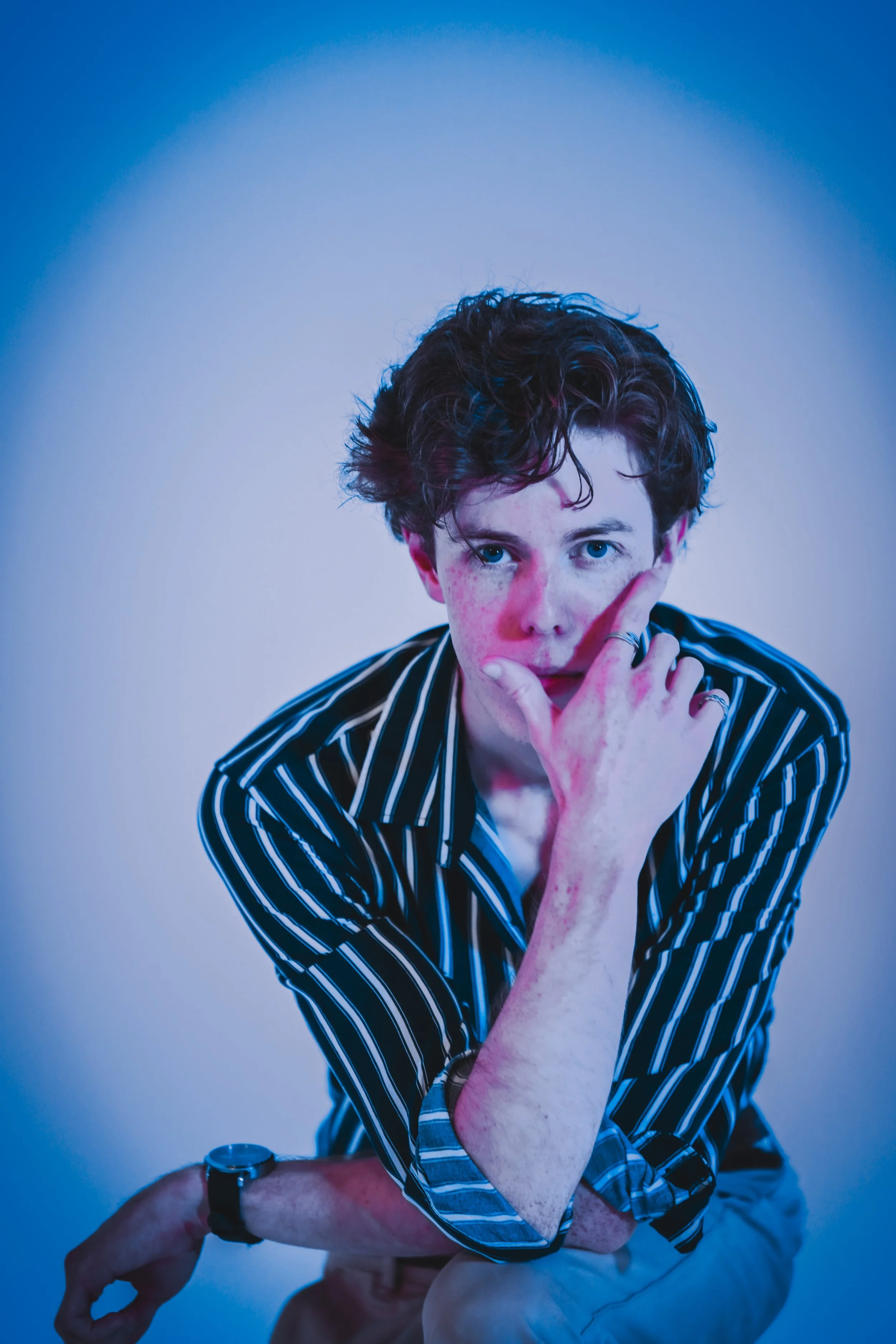 A young man with curly brown hair and blue eyes poses with his hand near his face, wearing a black and white striped shirt. The photo has vibrant blue and purple lighting with a plain light background.