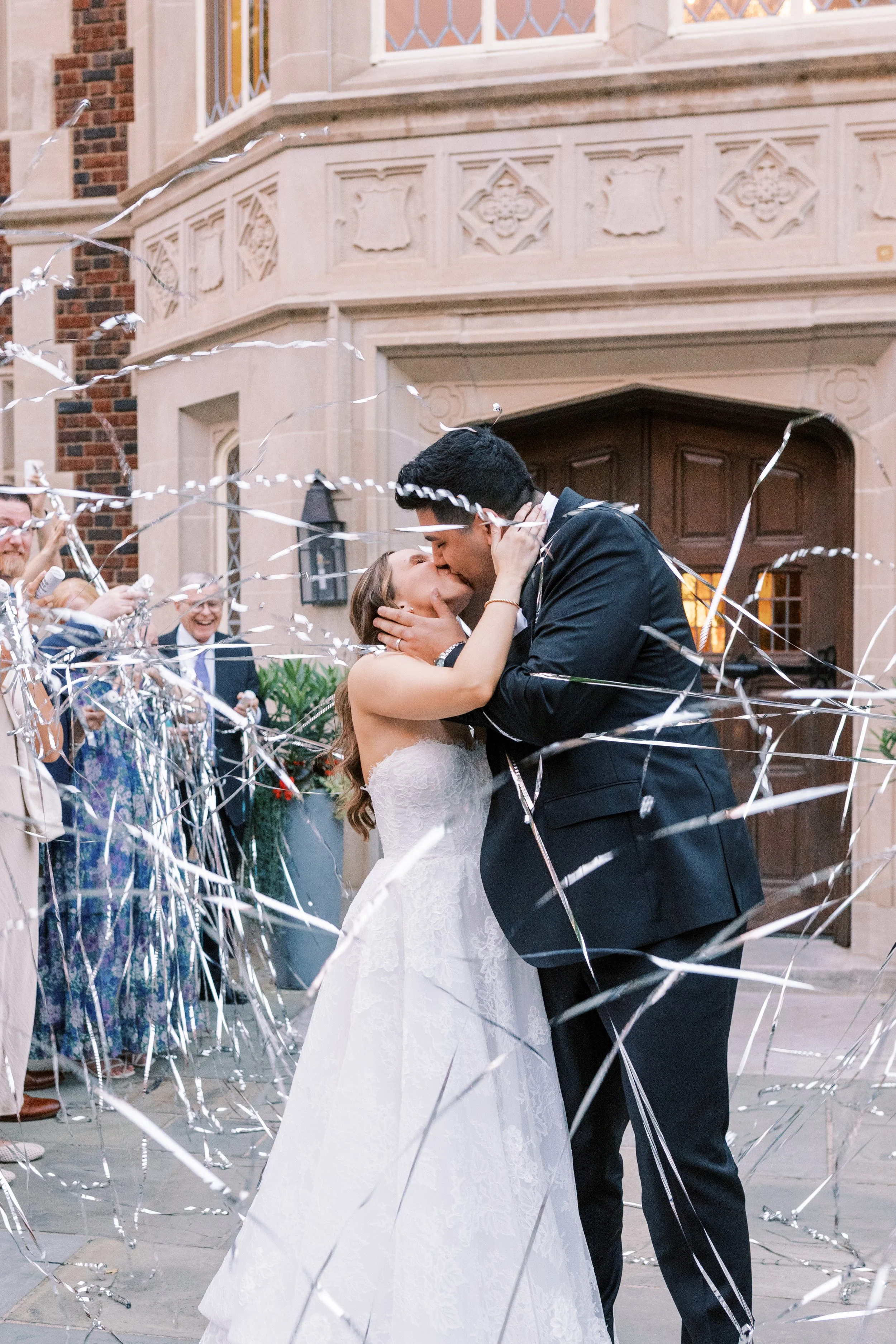 Bride and groom kiss surrounded by metallic streamers after wedding celebration outside a building with ornate architecture and friends celebrating.