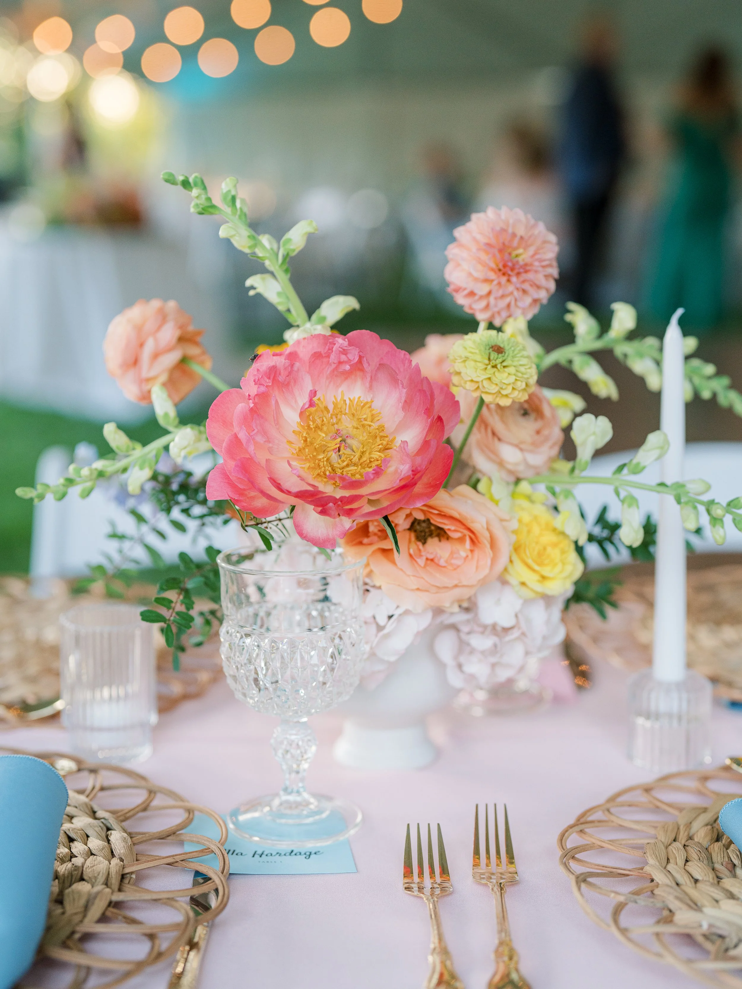 Bright pink and peach garden style centerpiece with textured greenery on outdoor wedding table with rattan chargers and blue napkins