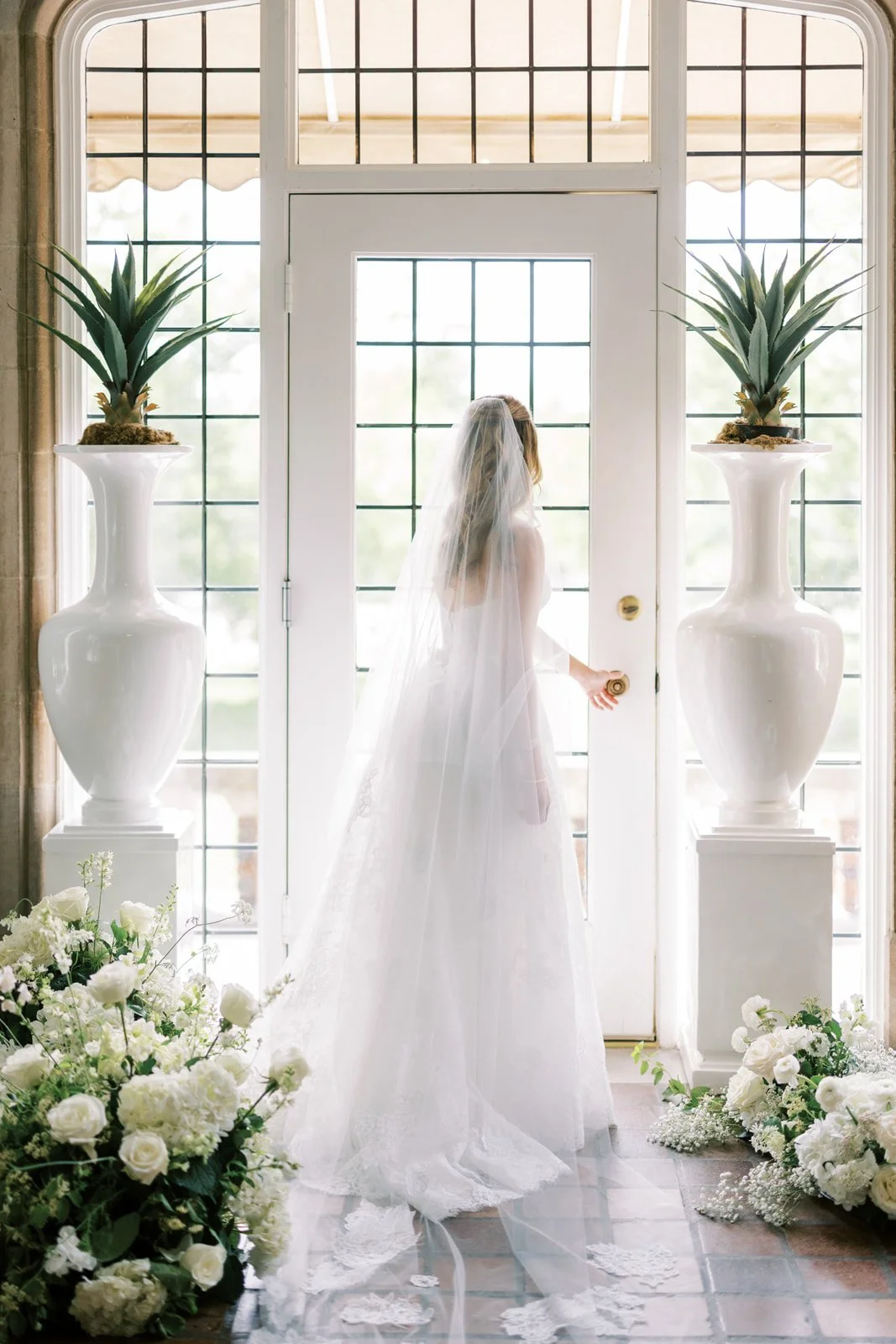 A bride in a wedding dress and veil, standing at a door with glass panels, flanked by large white vases with green plants, surrounded by white floral arrangements on the floor.