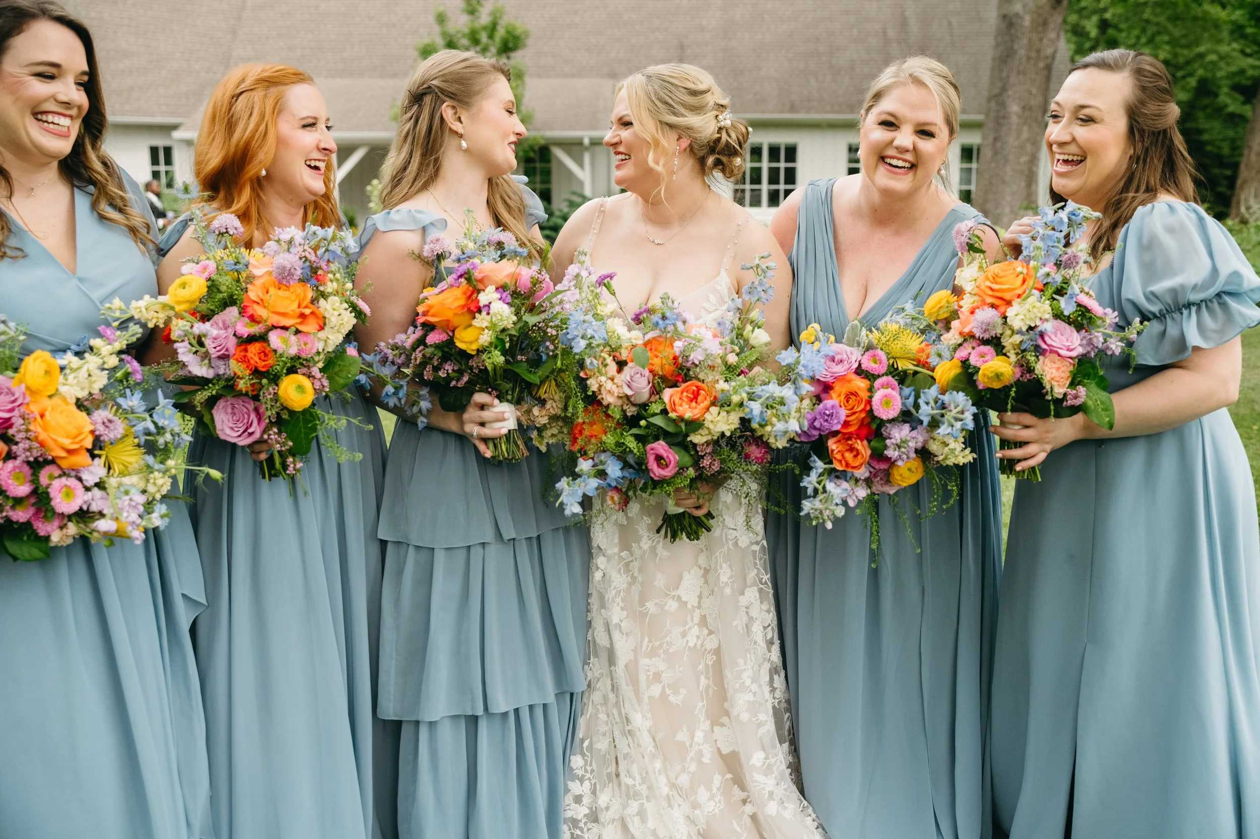 Bride and bridesmaids laughing together in soft blue dresses, holding vibrant spring bouquets with pops of pink, orange and yellow