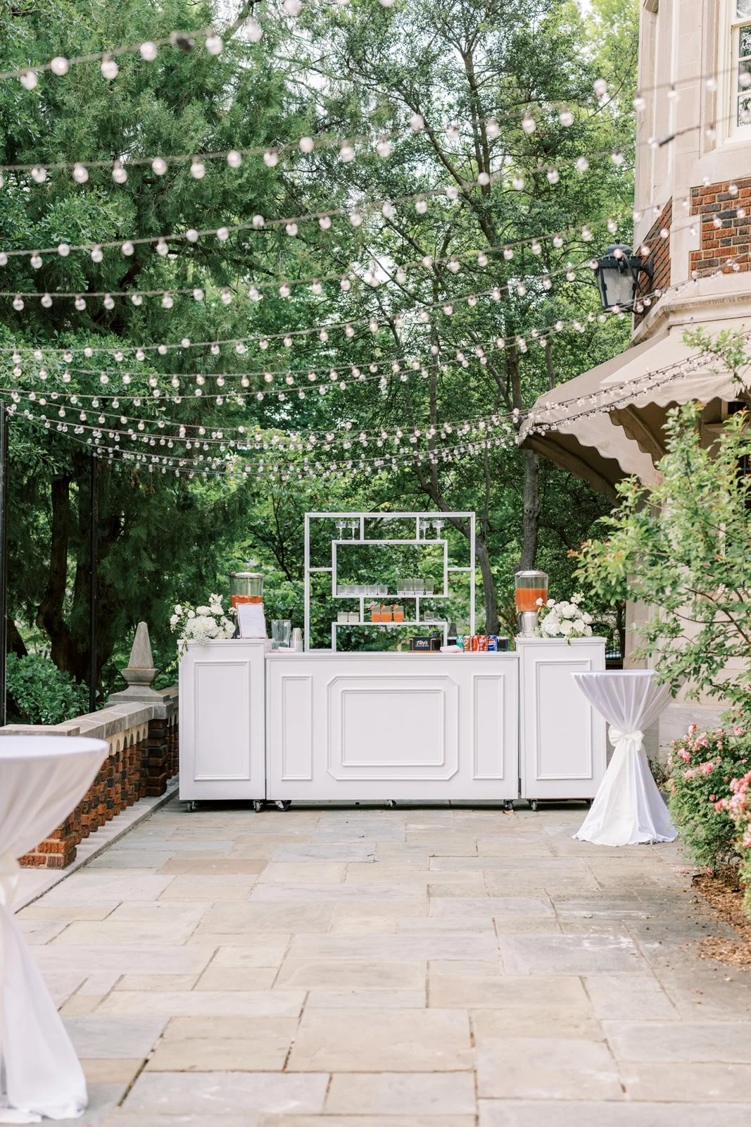 Outdoor event setup with string lights overhead, a white beverage bar, and two small round high-top tables with white tablecloths, in a lush garden with trees and flowers.