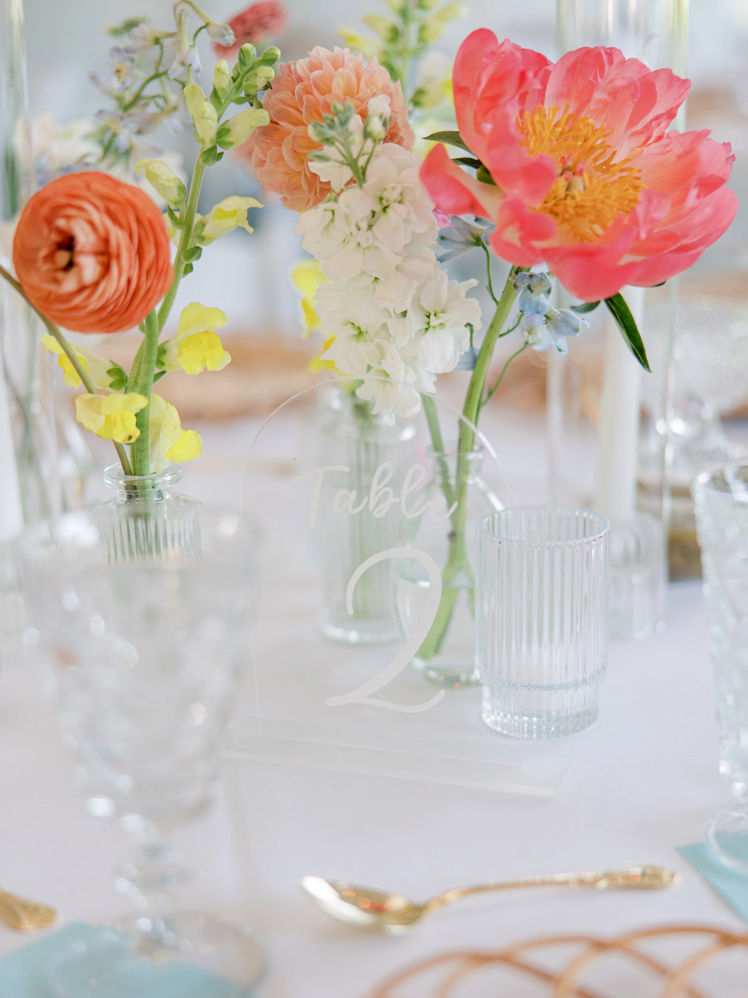 Bright coral and pink peonies with ranunculus and yellow flower accents in bud vases on reception table