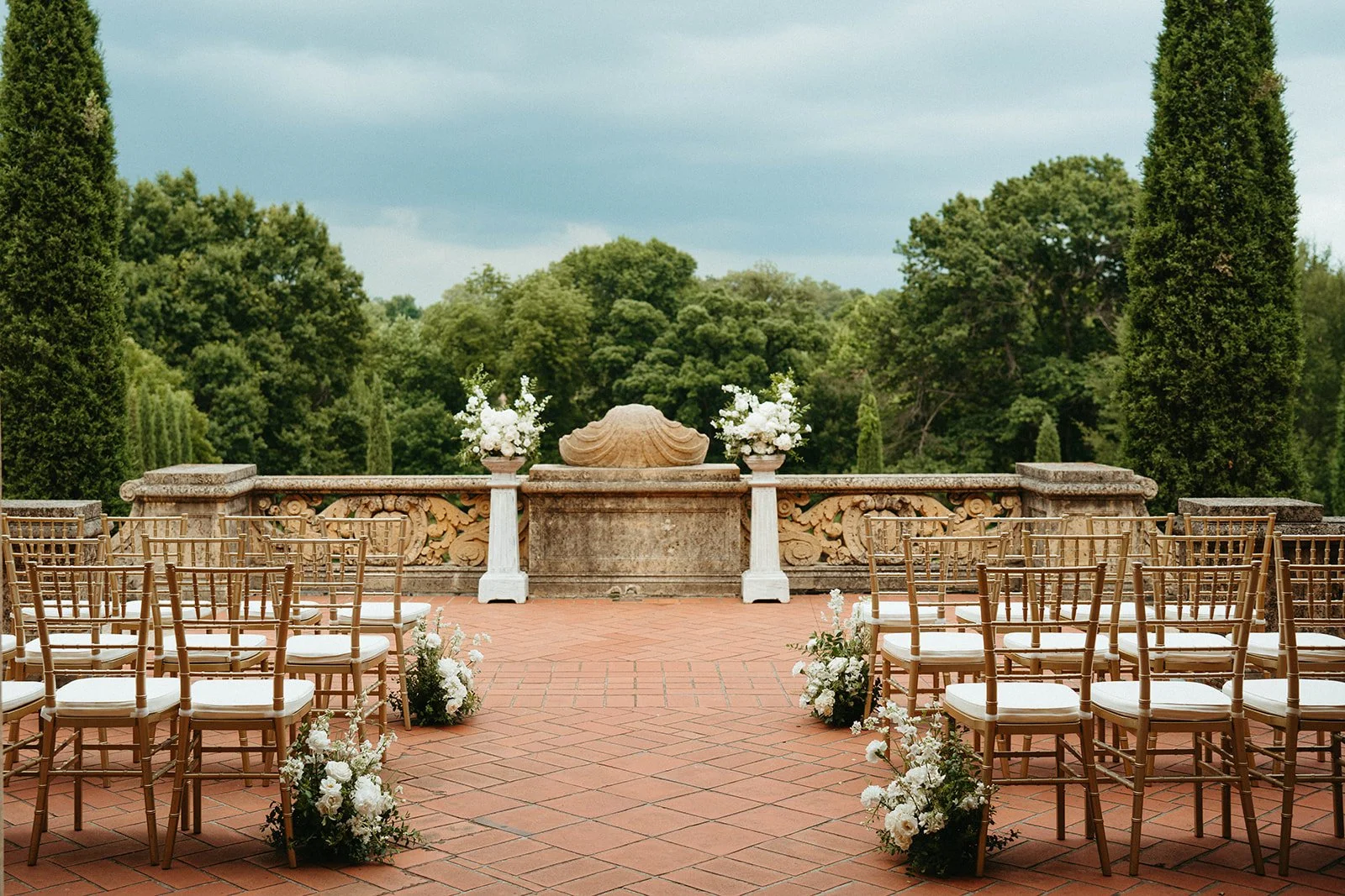 Outdoor wedding ceremony setup with rows of gold chairs and floral arrangements, on a brick patio with a stone balustrade and lush green trees in the background.
