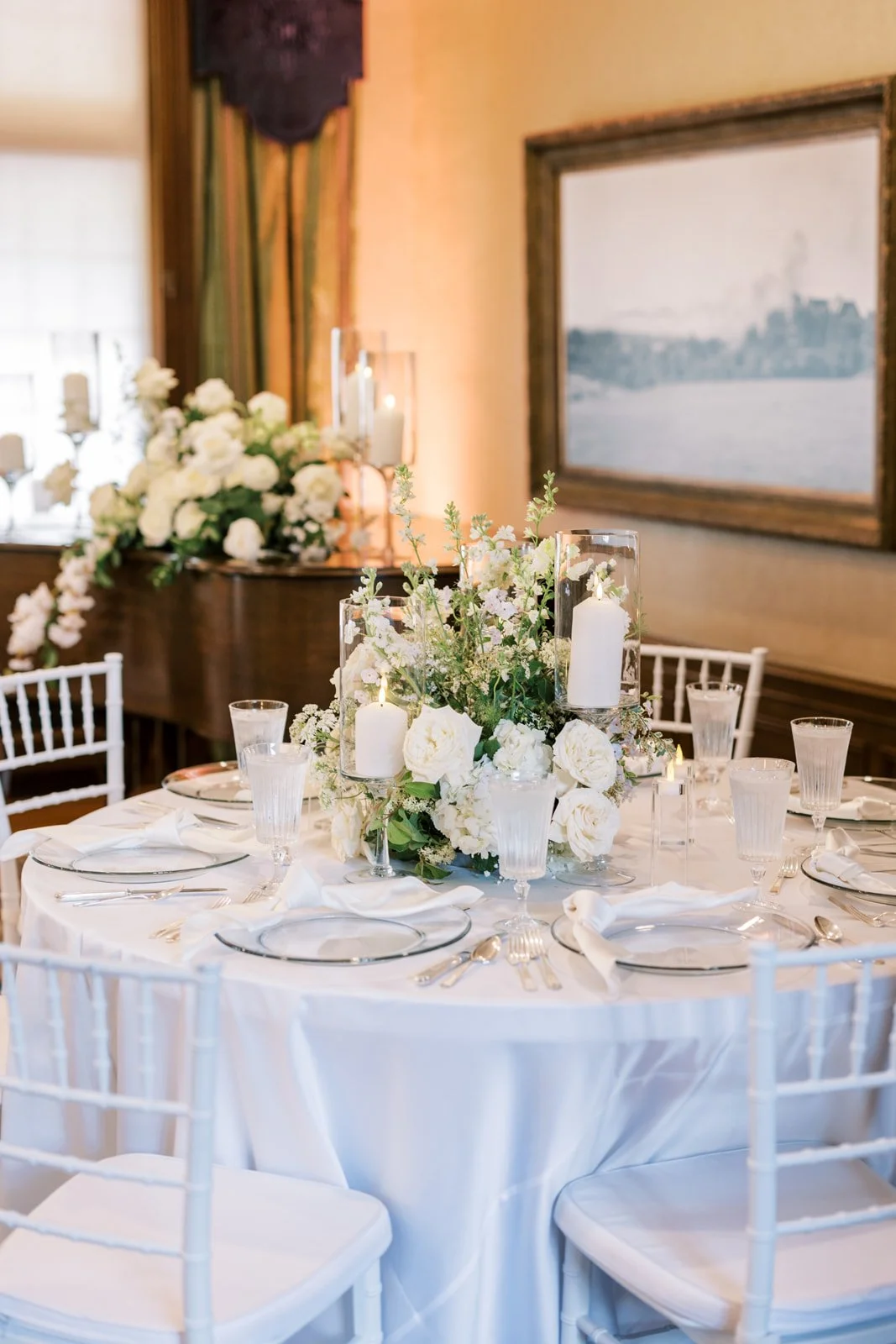 Elegant dining table set for a wedding or special event with a white tablecloth, white floral centerpiece, candles, and glassware in a decorated banquet room.