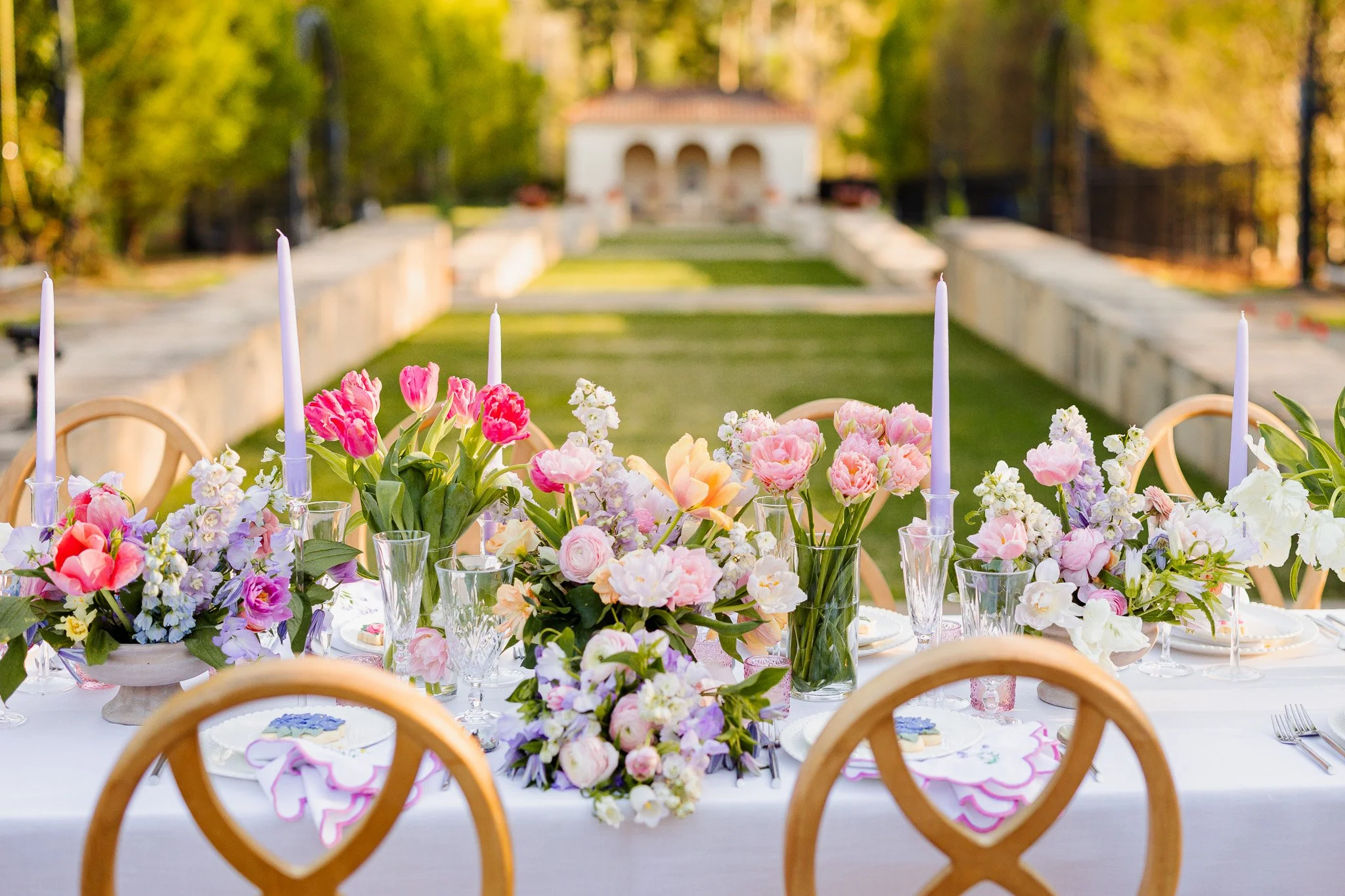 Romantic outdoor wedding table with pastel floral arrangements, natural wood chairs, and garden venue backdrop