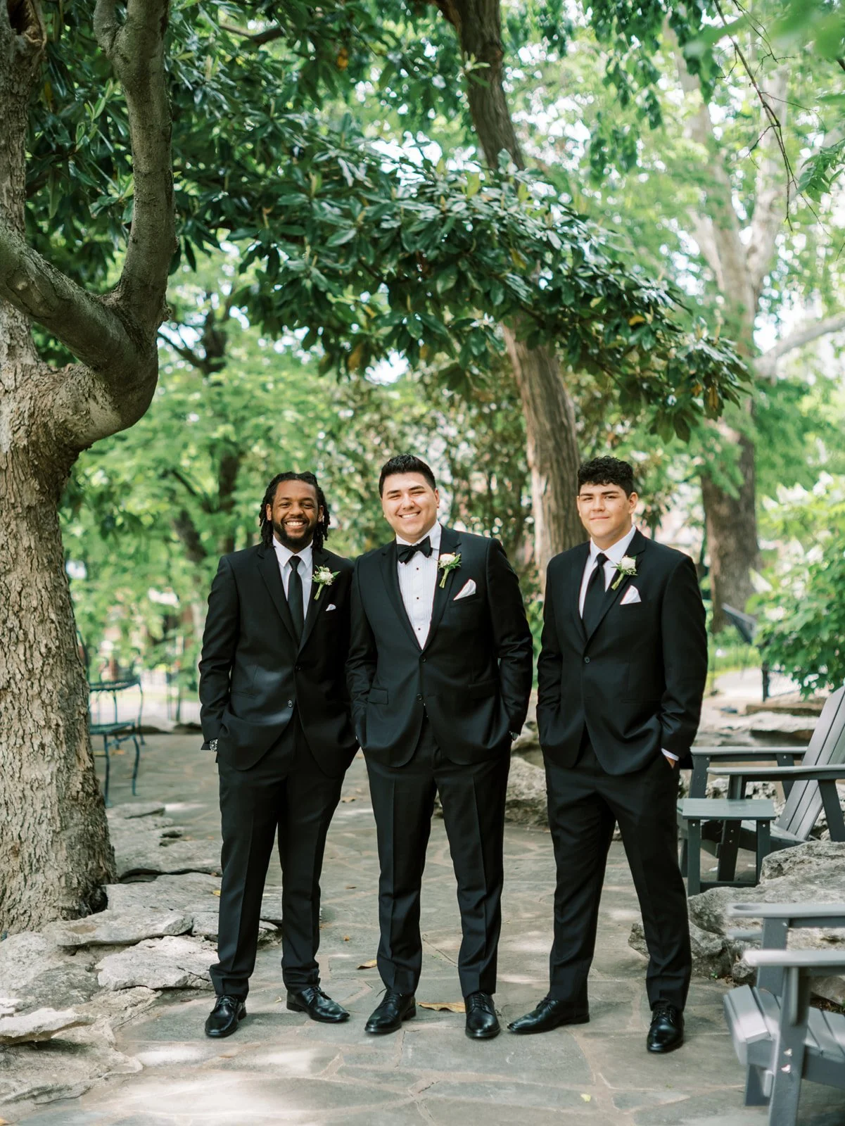 Three men in black tuxedos with white shirts, black ties, and boutonnières, standing outdoors on a stone pathway with trees and greenery in the background.