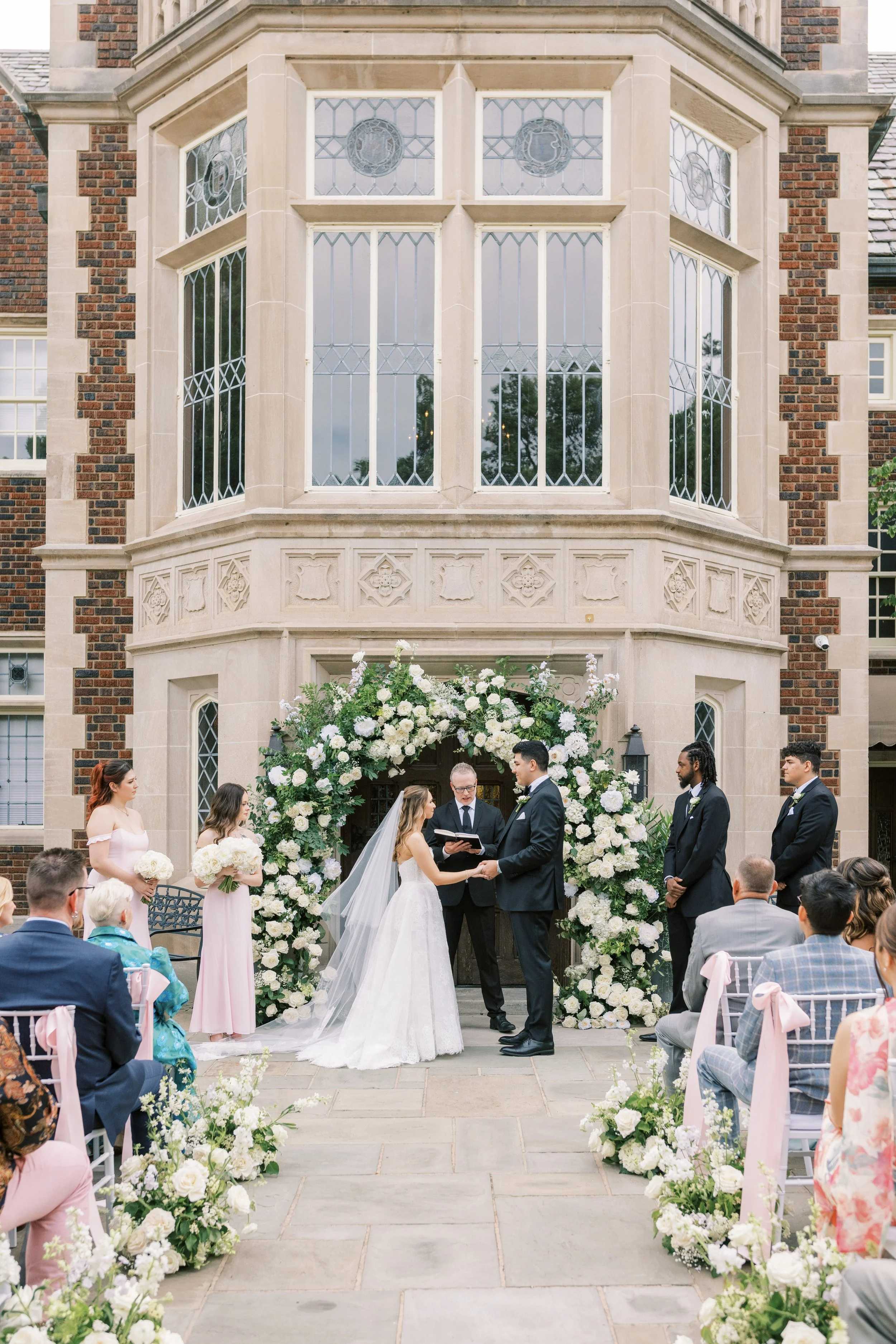 A wedding ceremony taking place outdoors in front of a large building with ornate windows. The bride and groom are holding hands and facing each other under a floral arch. Two women are standing on the left holding bouquets, and four men on the right are dressed in suits. Guests are seated on either side, and the setting is decorated with flowers and pink ribbons.